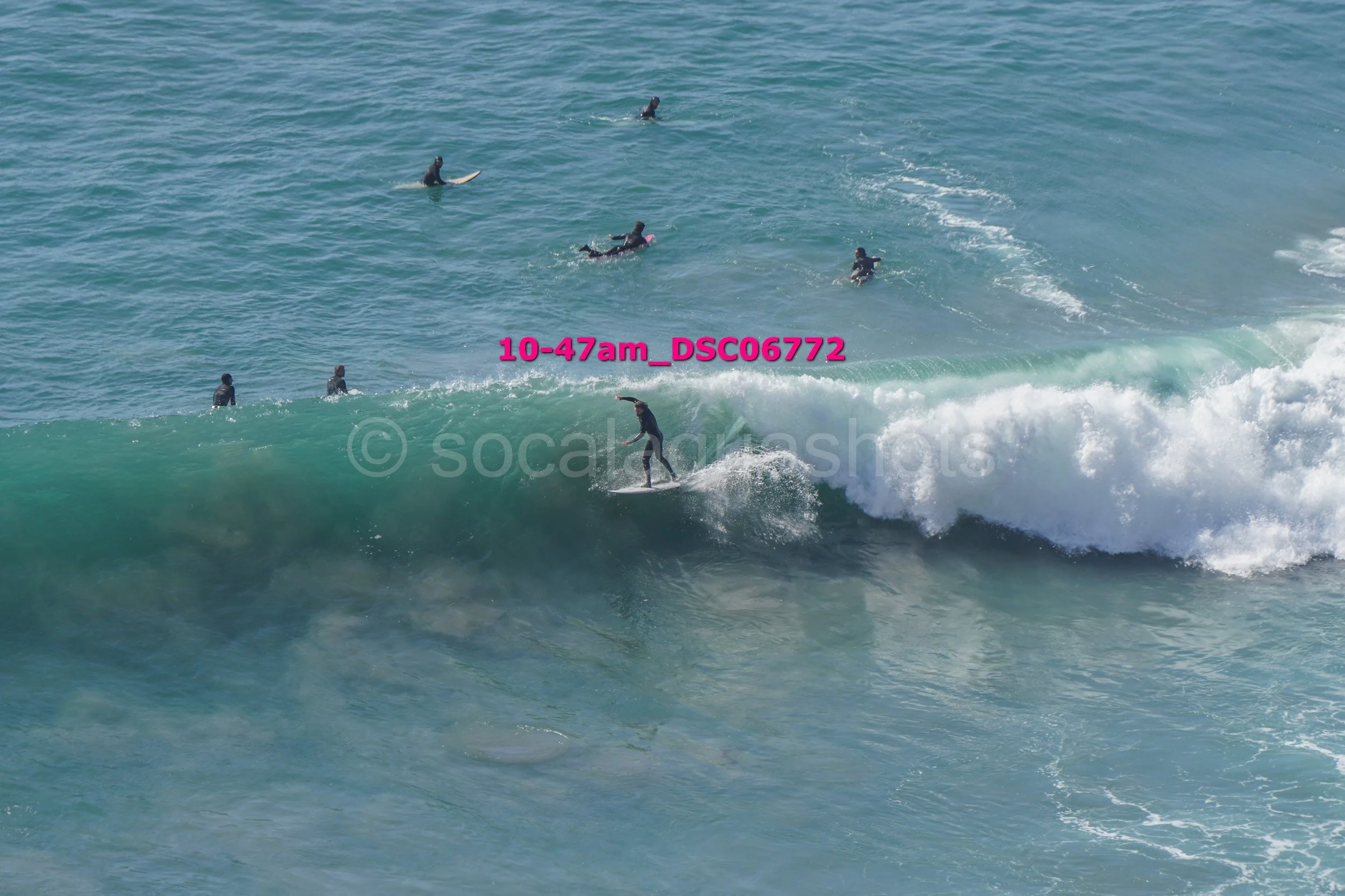 Surfer riding a wave while ten people in wetsuits watch from the water.