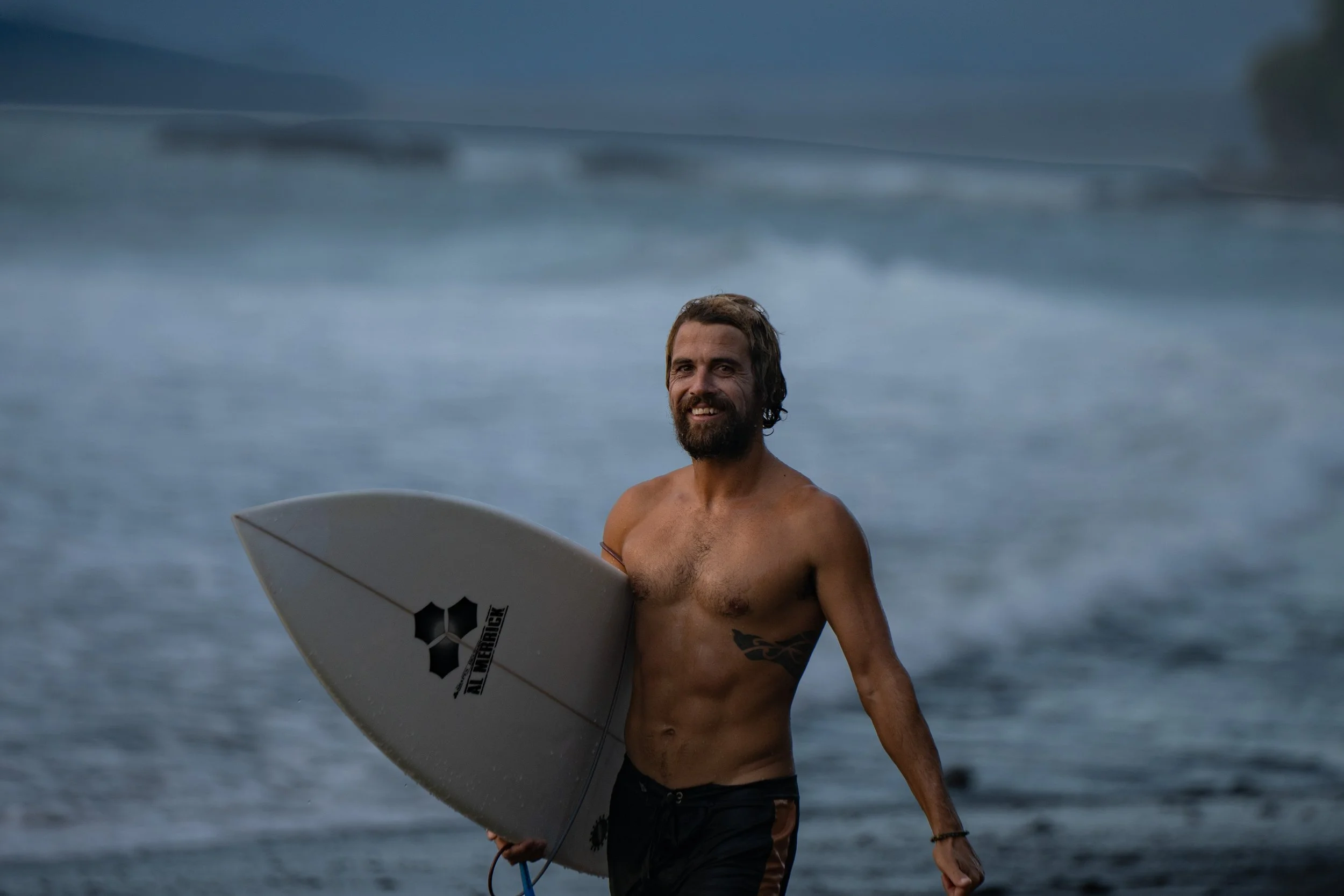 A shirtless man with a beard and tattoos holding a surfboard, standing on the beach with the ocean and cloudy sky in the background.