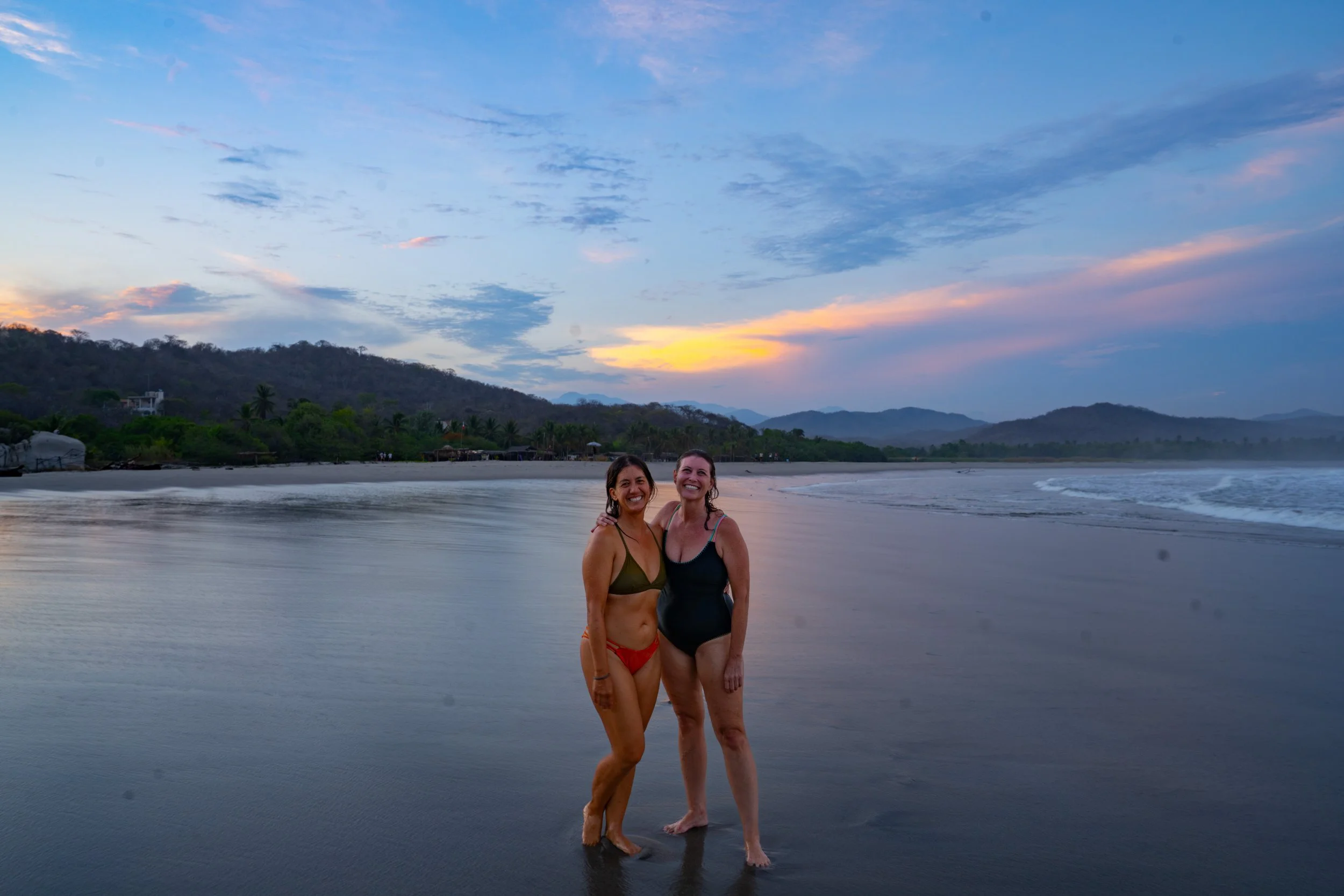 Two women in swimsuits standing on a beach during sunset with a mountain landscape and a partly cloudy sky in the background.