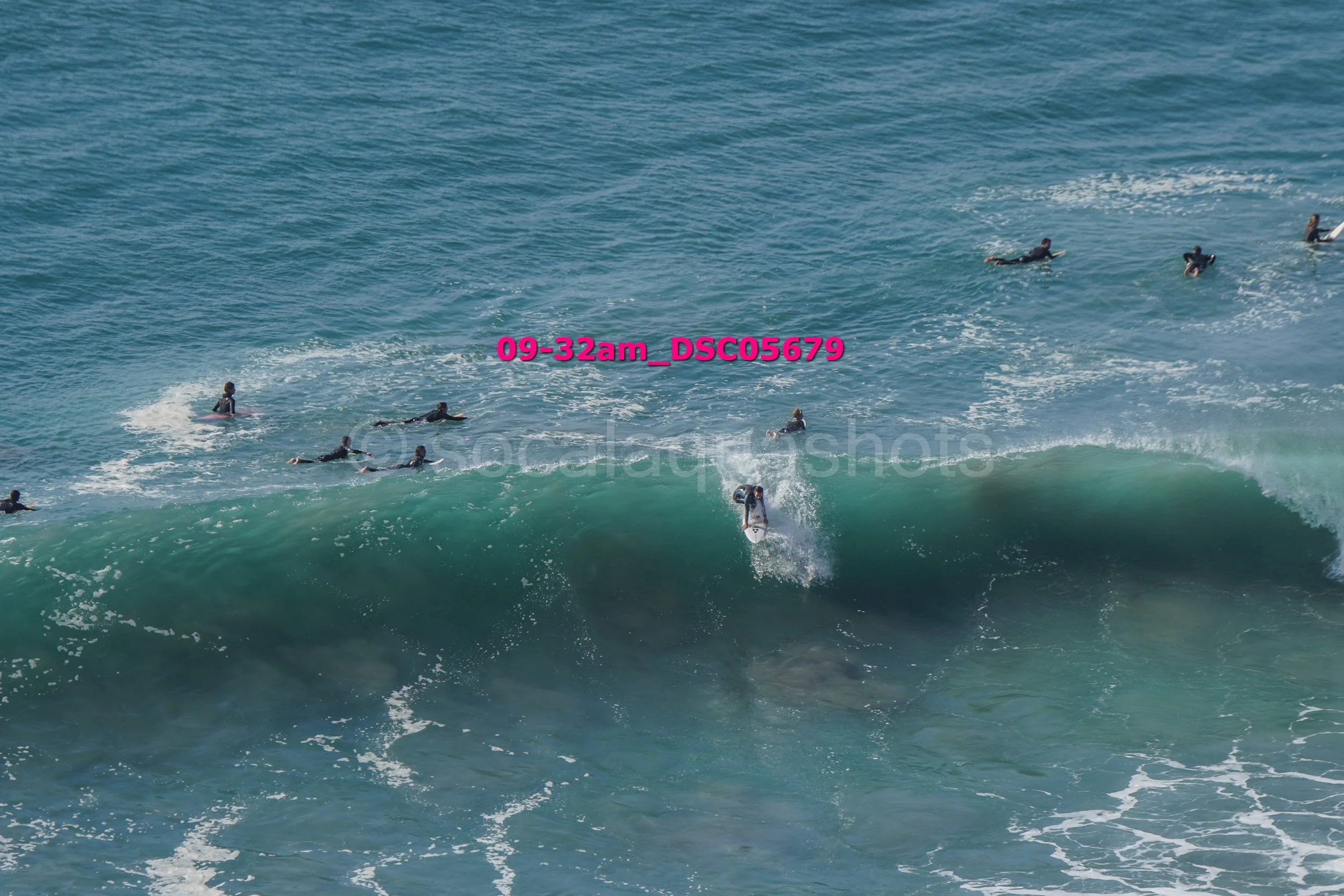 A group of surfers in the ocean, with one caught riding a wave while others wait or paddle nearby.