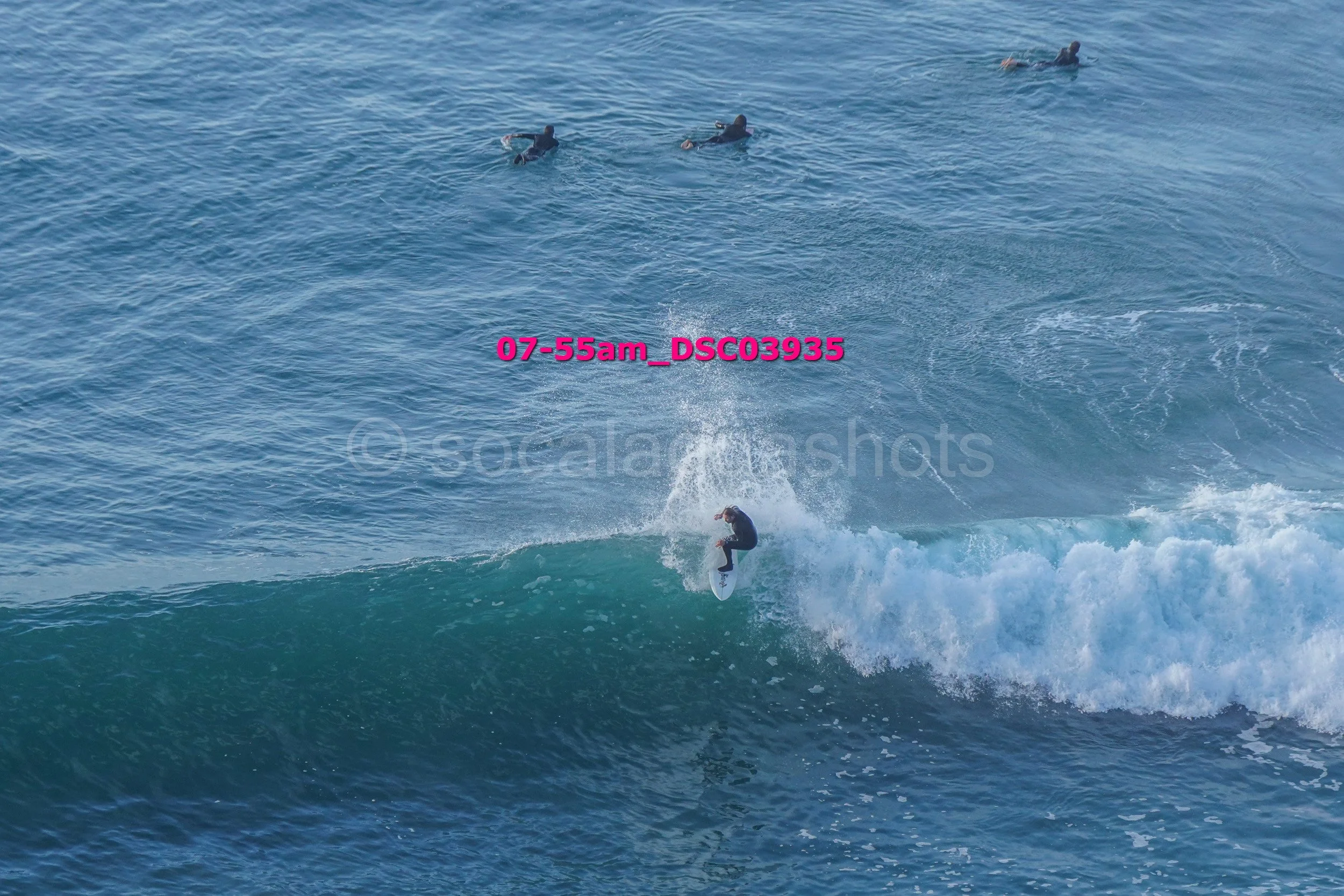 A surfer riding a wave in the ocean with three other people swimming or floating in the water nearby.