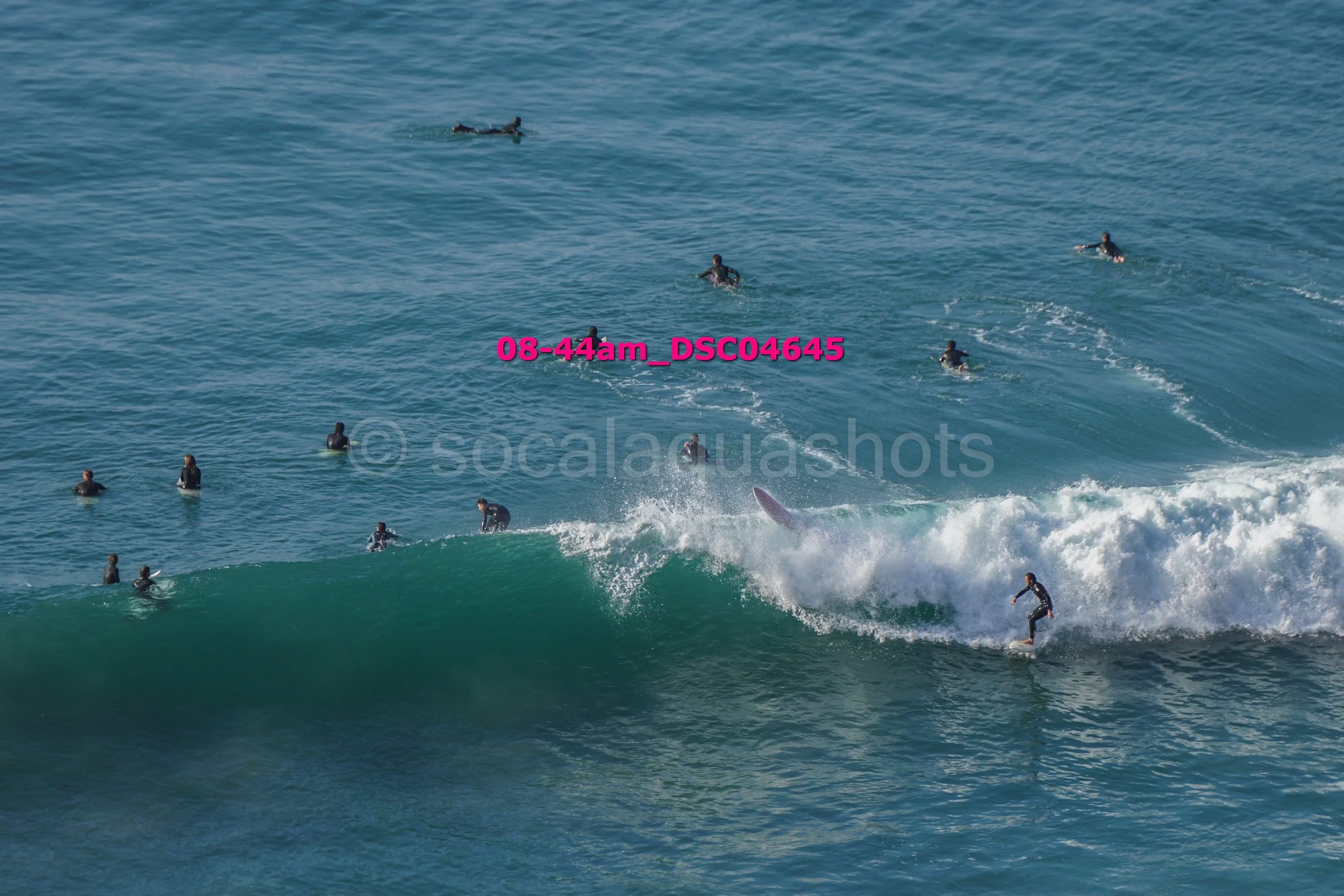 Surfers riding and waiting for waves in the ocean, some on surfboards and some in the water, with a large wave in the foreground.