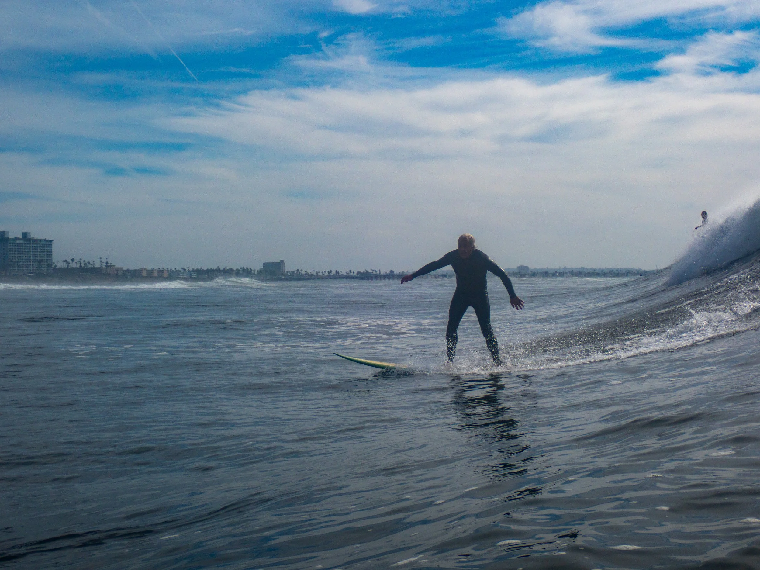 A person surfing on a wave in the ocean with a city skyline and palm trees in the background, and a blue sky with some clouds.