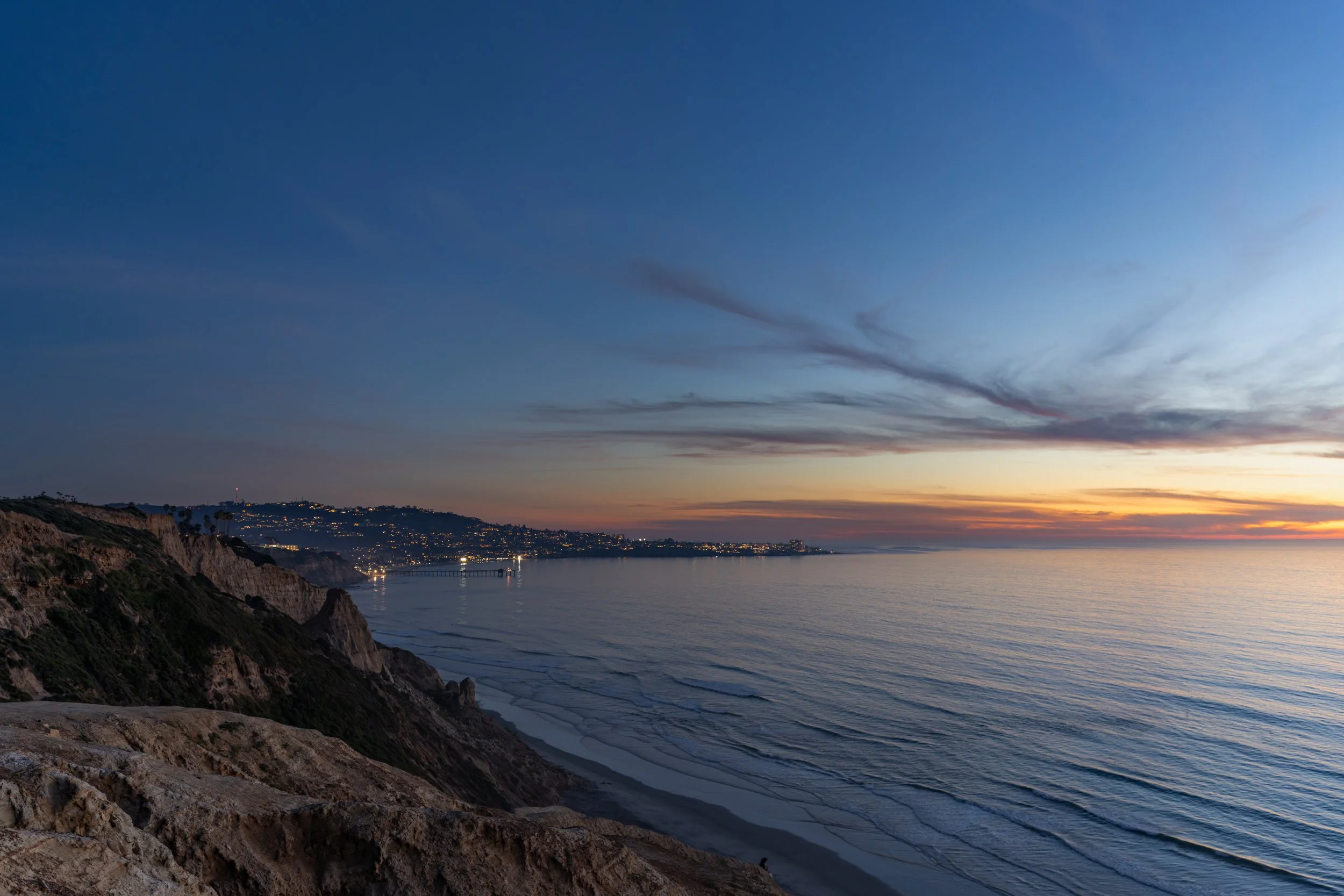 Sunset over the ocean with cliffs in the foreground, a distant cityscape, and a pier extending into the water.