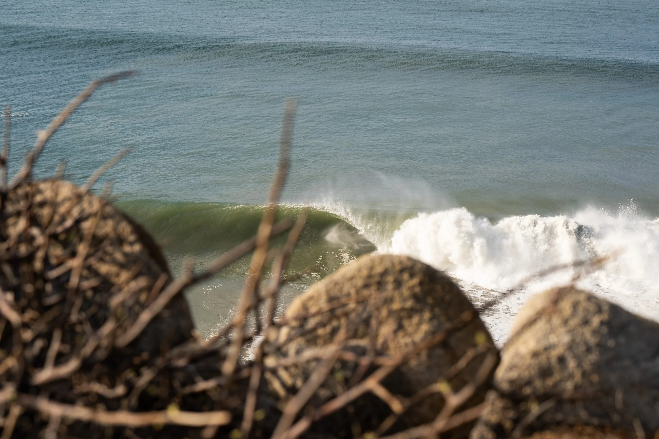 View of the ocean with waves crashing on the shore, seen through rocks and dried branches in the foreground.