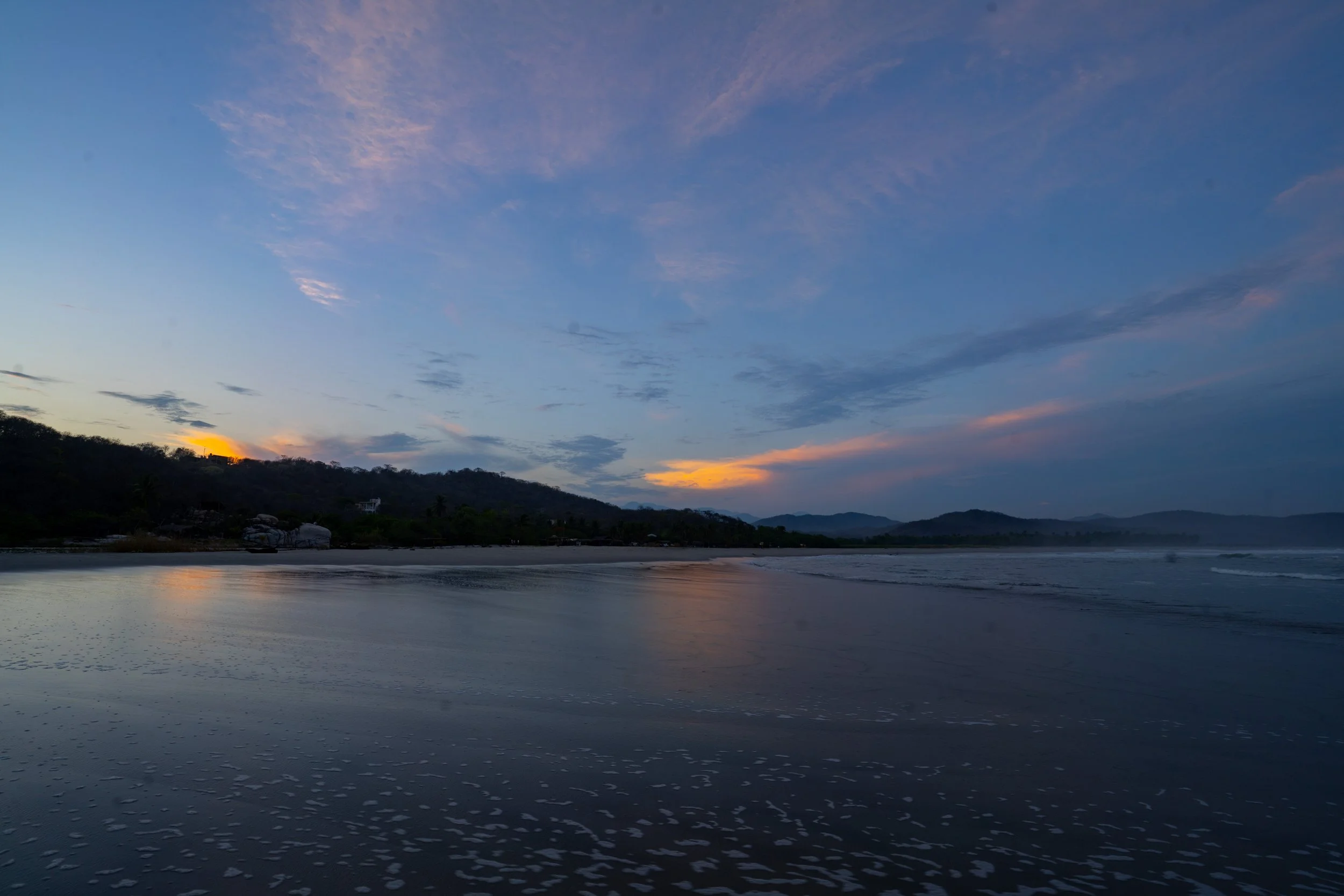 Sunset over a beach with calm water, silhouettes of mountains and trees in the background, and colorful sky with pink and orange hues