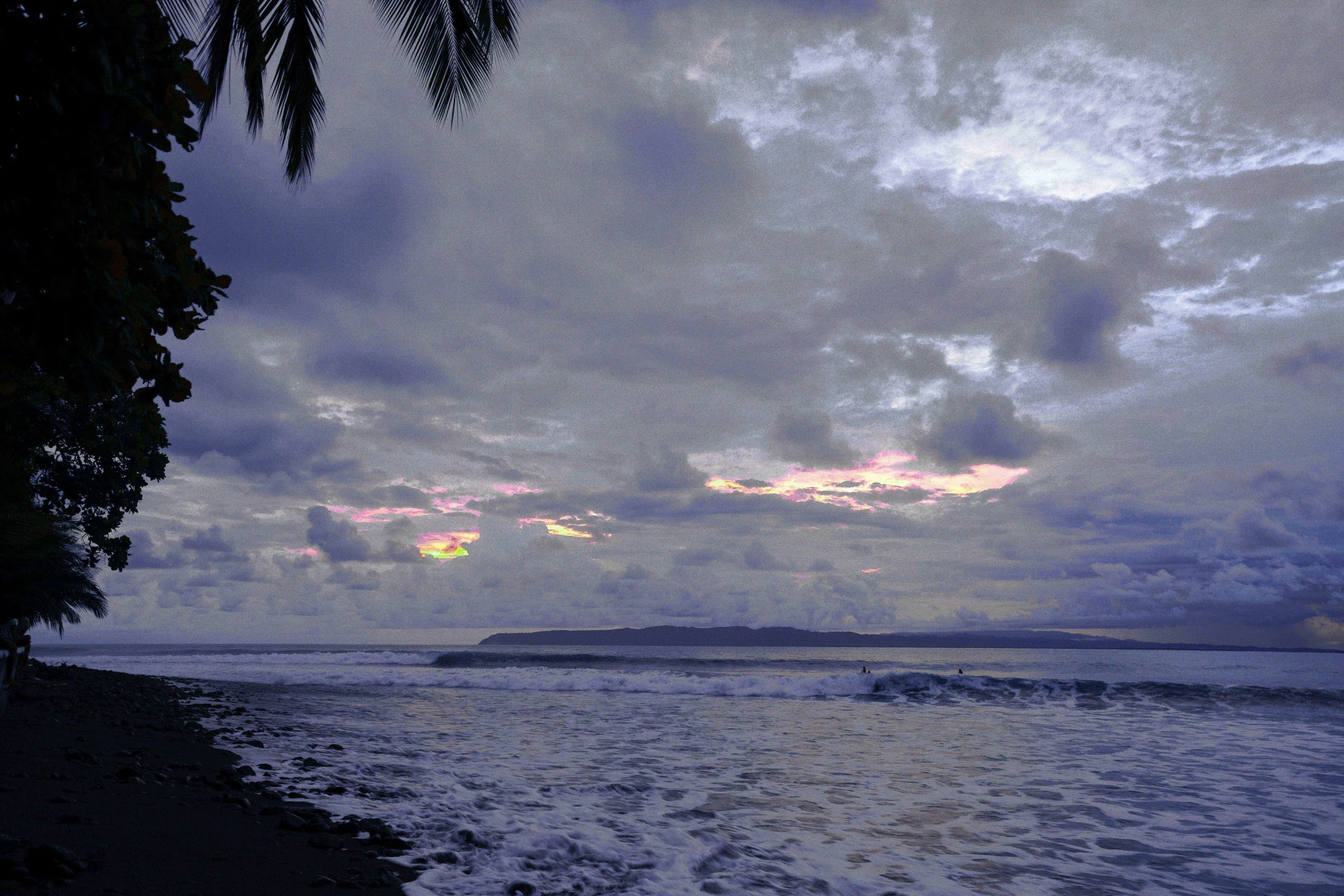 A coastal scene during dusk with cloudy sky, a hint of pink and purple hues, palm trees on the left, and a distant landmass across the ocean.