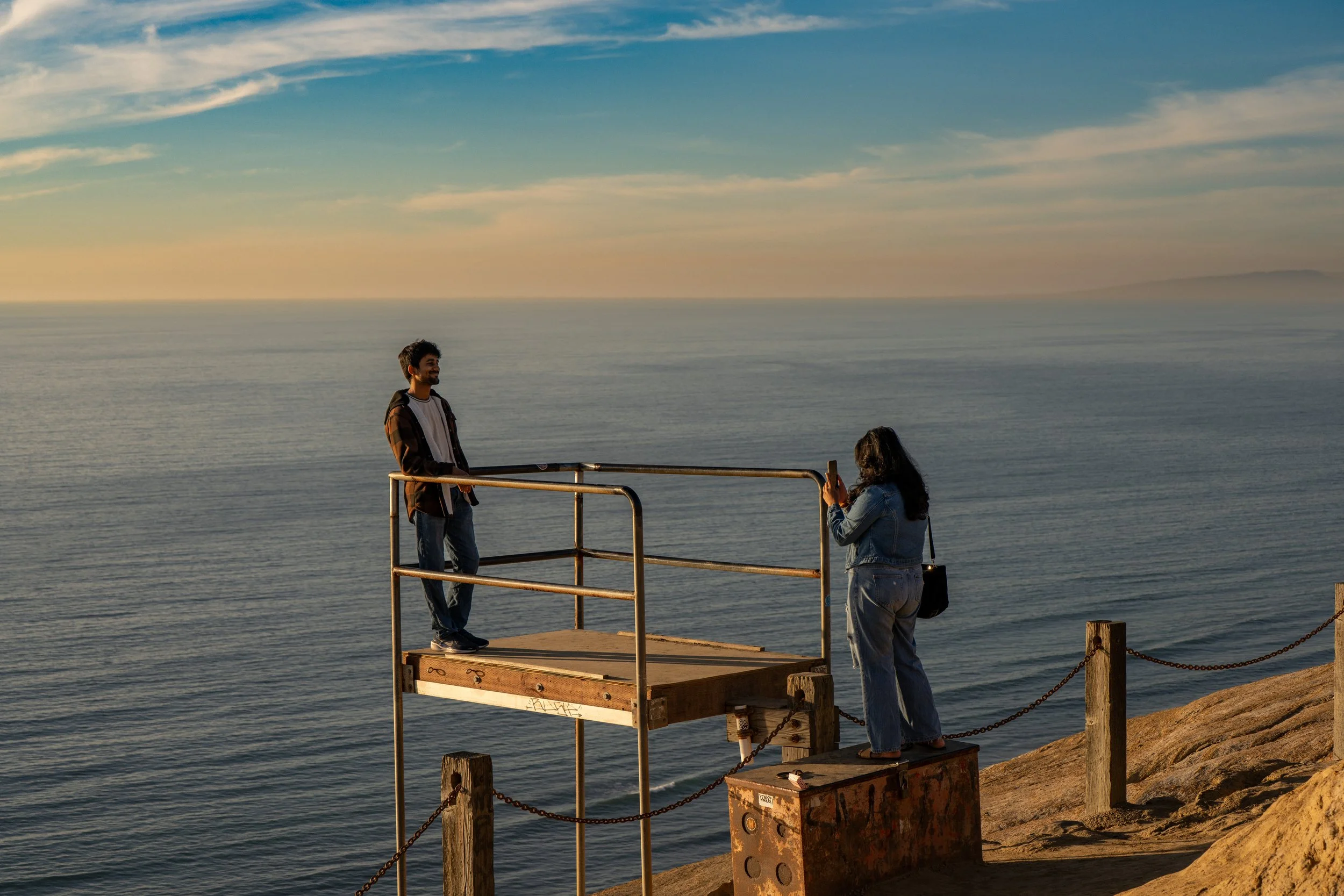 A man and a woman are standing on a wooden platform near the ocean at sunset, with the man smiling and the woman taking a photo of him.