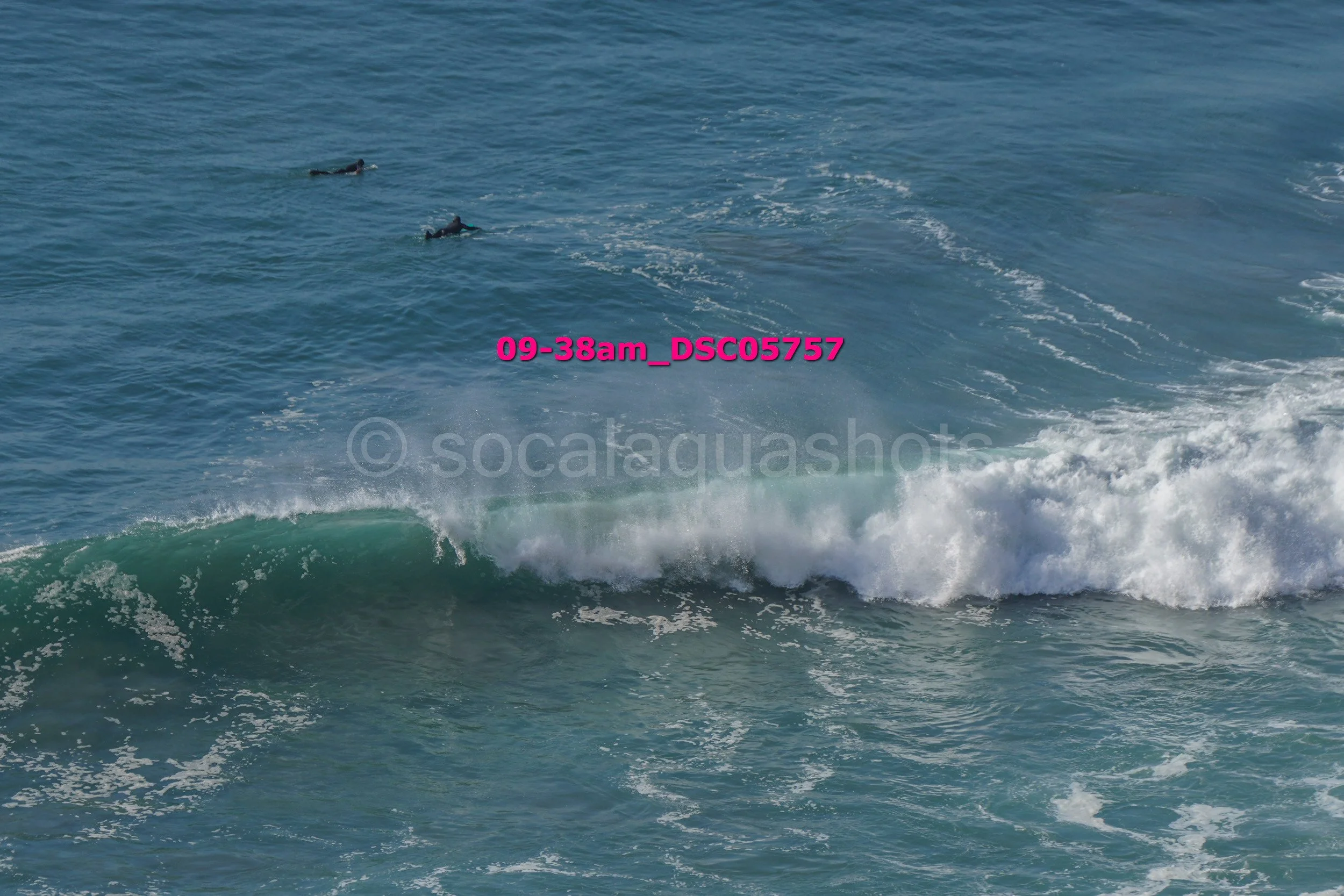 Two surfers lying on their surfboards in the ocean, with a breaking wave in the foreground.