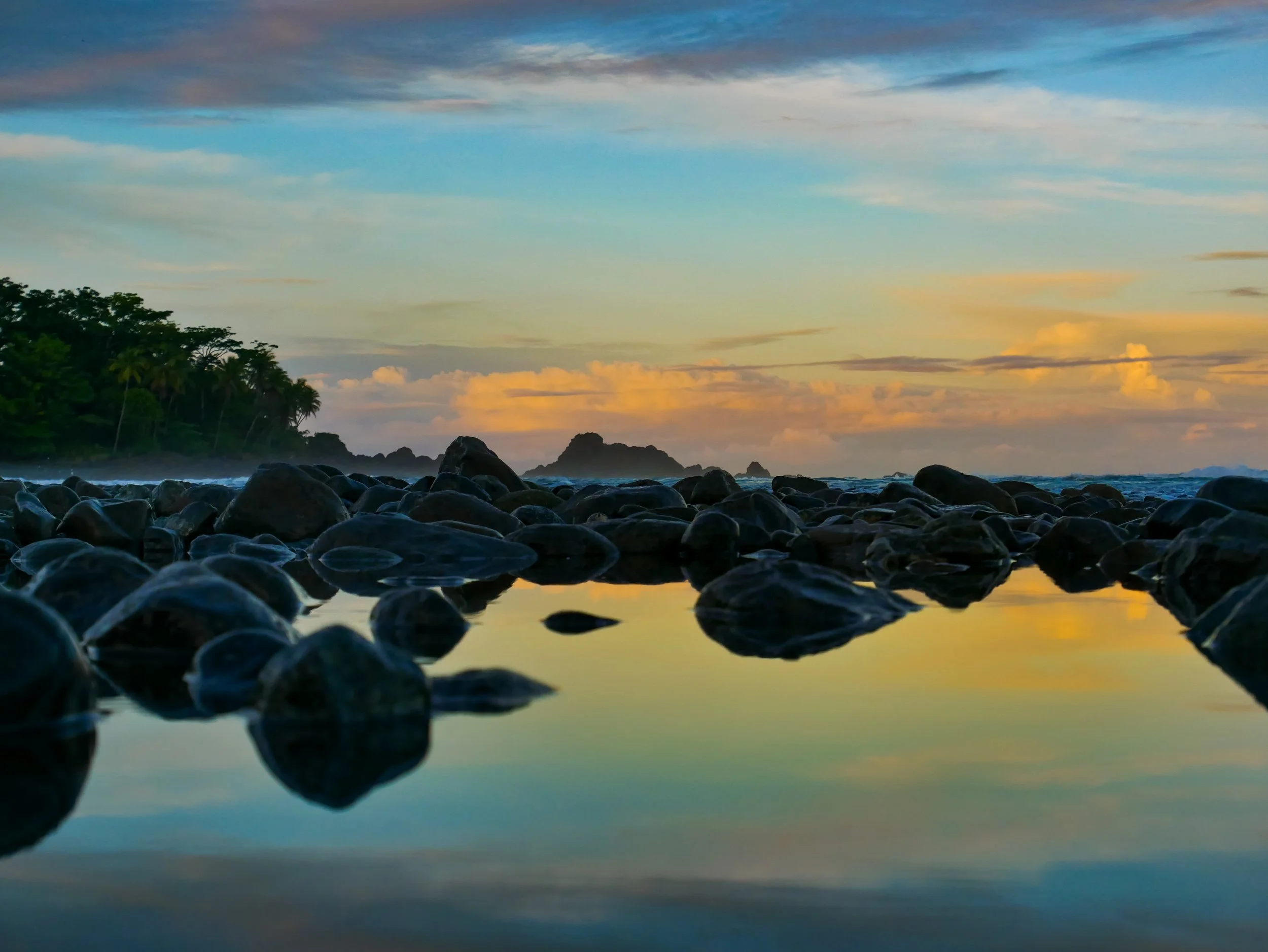 Rocky beach with calm water reflecting the sky and a distant tree-covered coastline at sunset with orange, yellow, and blue clouds.