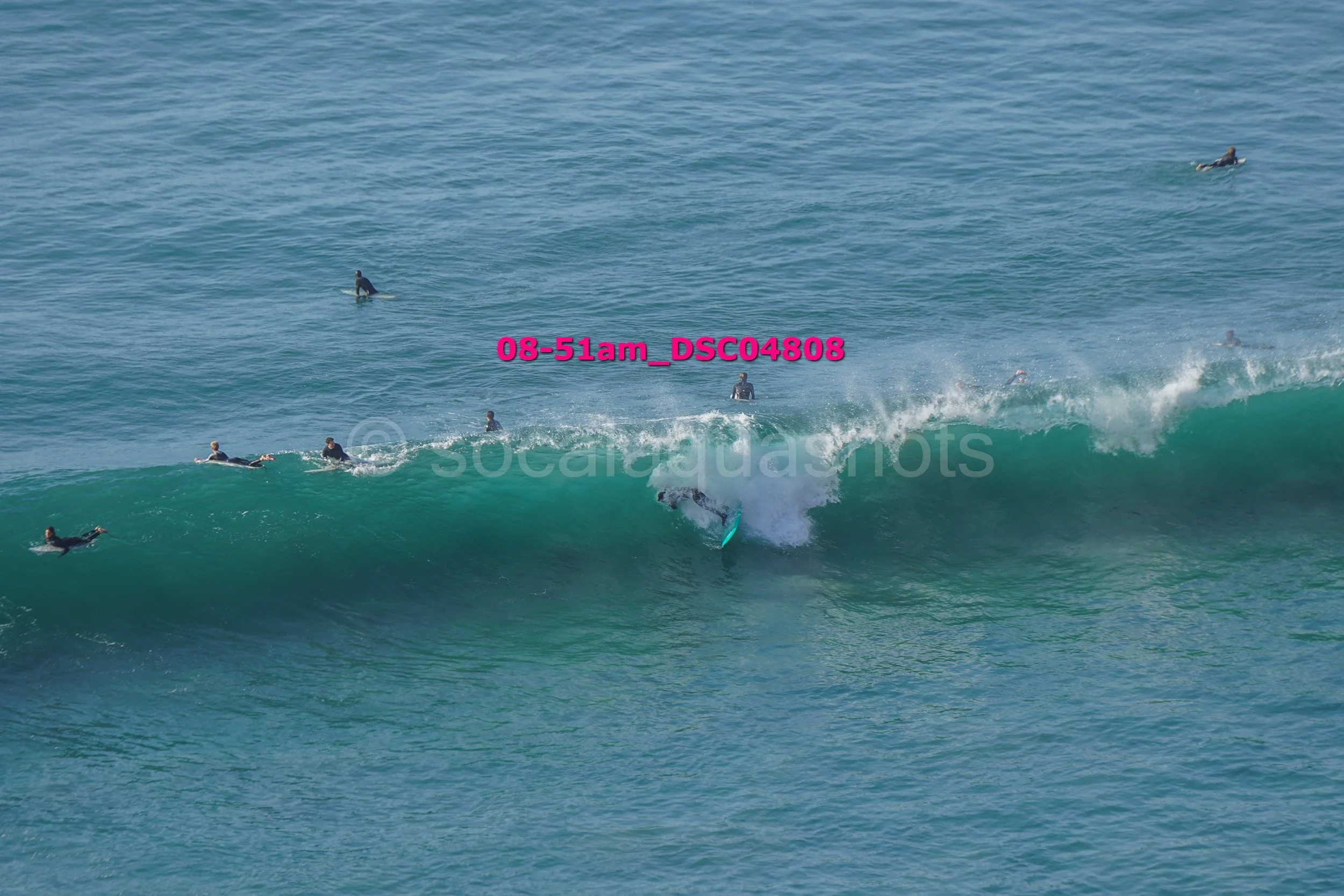 Surfer riding a wave with several people in wetsuits in the water around, some paddling and some sitting on surfboards, in an ocean scene.
