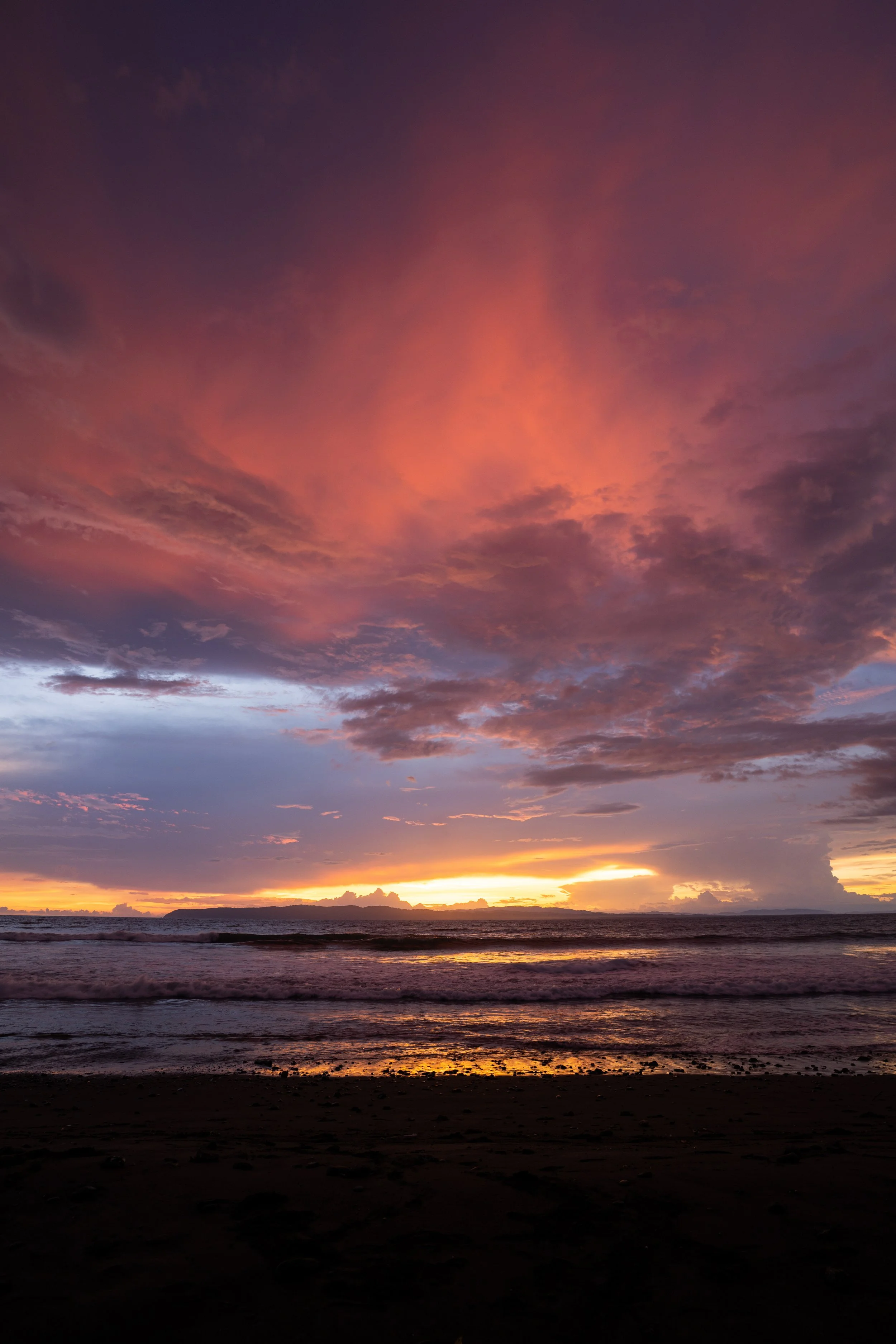 Sunset over the ocean with dark clouds, pink, orange, and purple hues in the sky, and waves gently crashing on the beach.