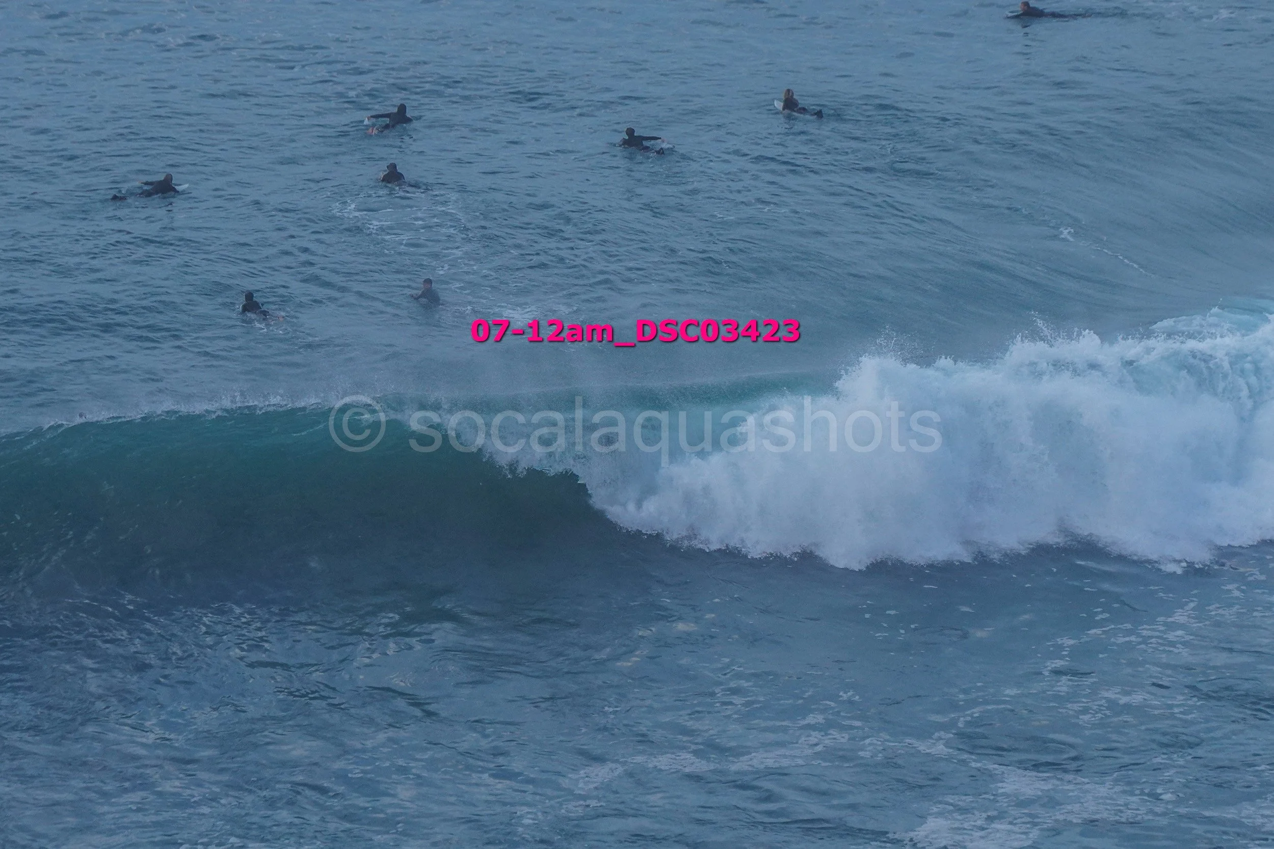 Several surfers in the ocean waiting near a breaking wave with text overlay '07-12am_DSC3423' in pink.