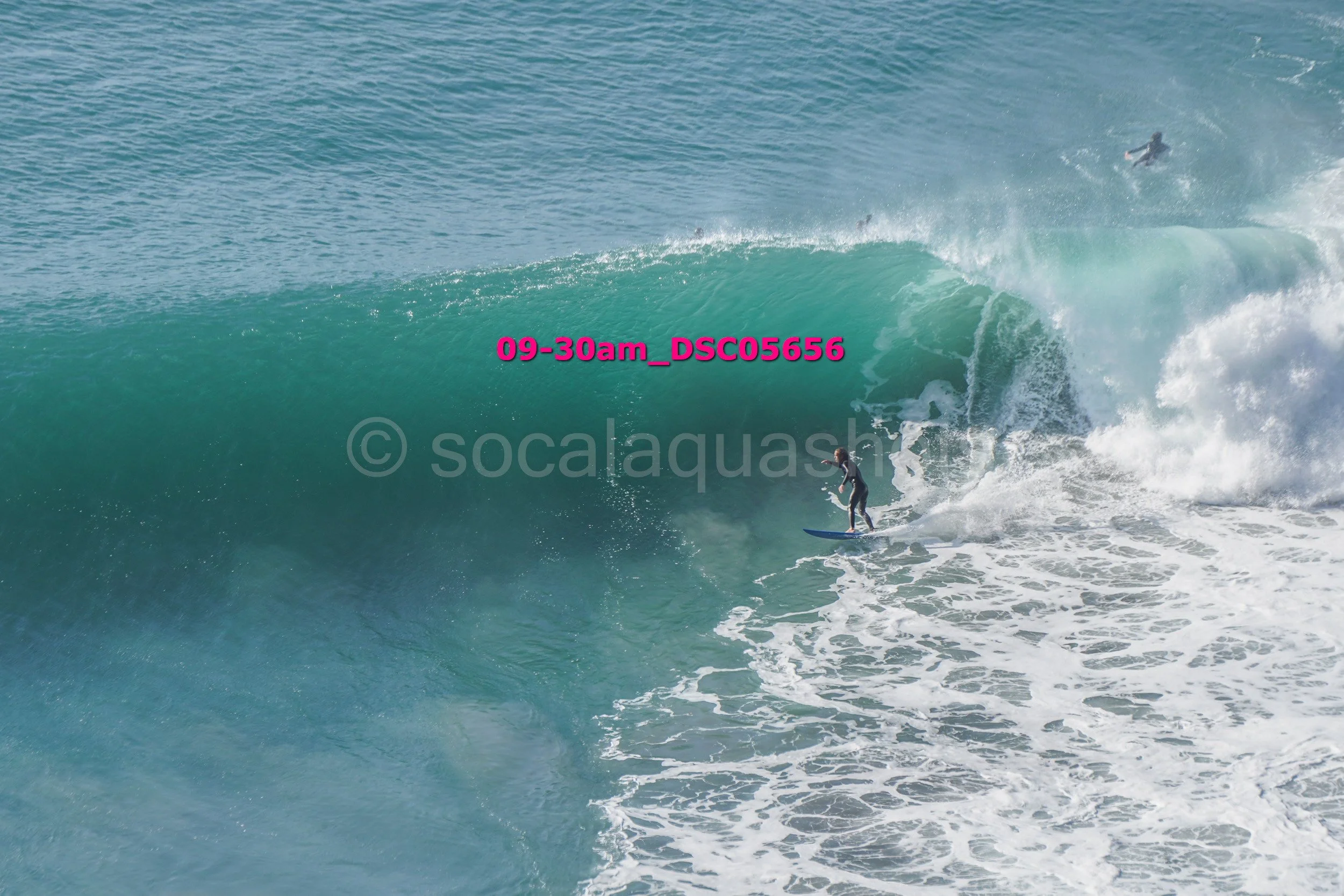 Surfer riding a large ocean wave, with another surfer visible in the water in the background.