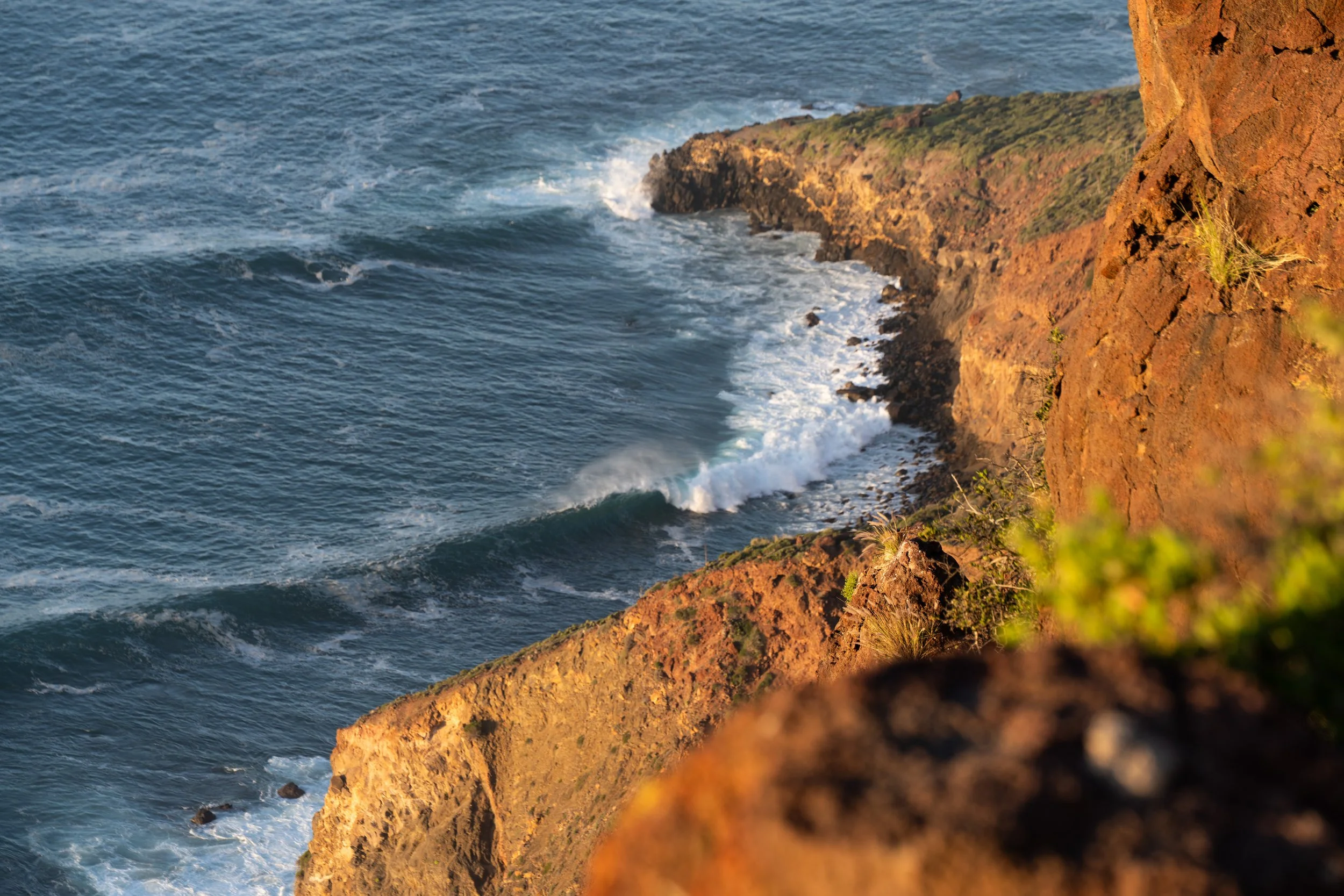 Ocean waves crashing against reddish-brown rocky cliffs under sunlight.