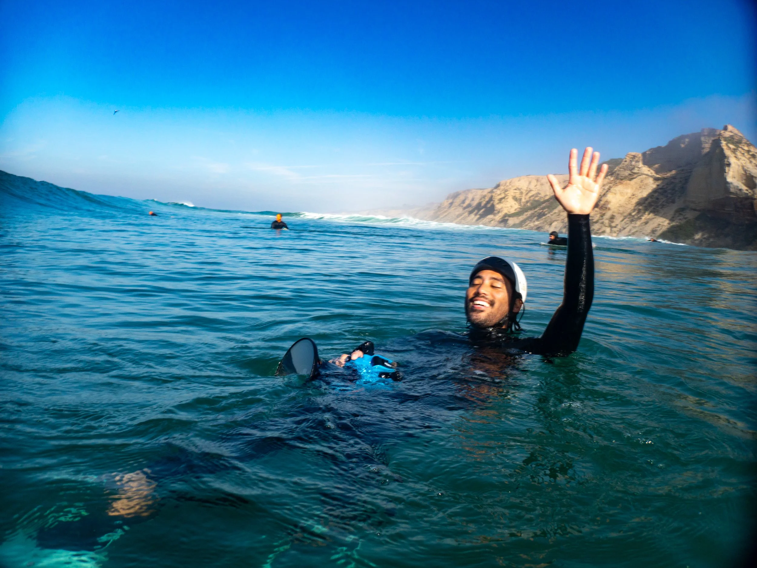 Person in a wetsuit and helmet floating in the ocean, smiling and waving, with rocky cliffs in the background and other people in the water.