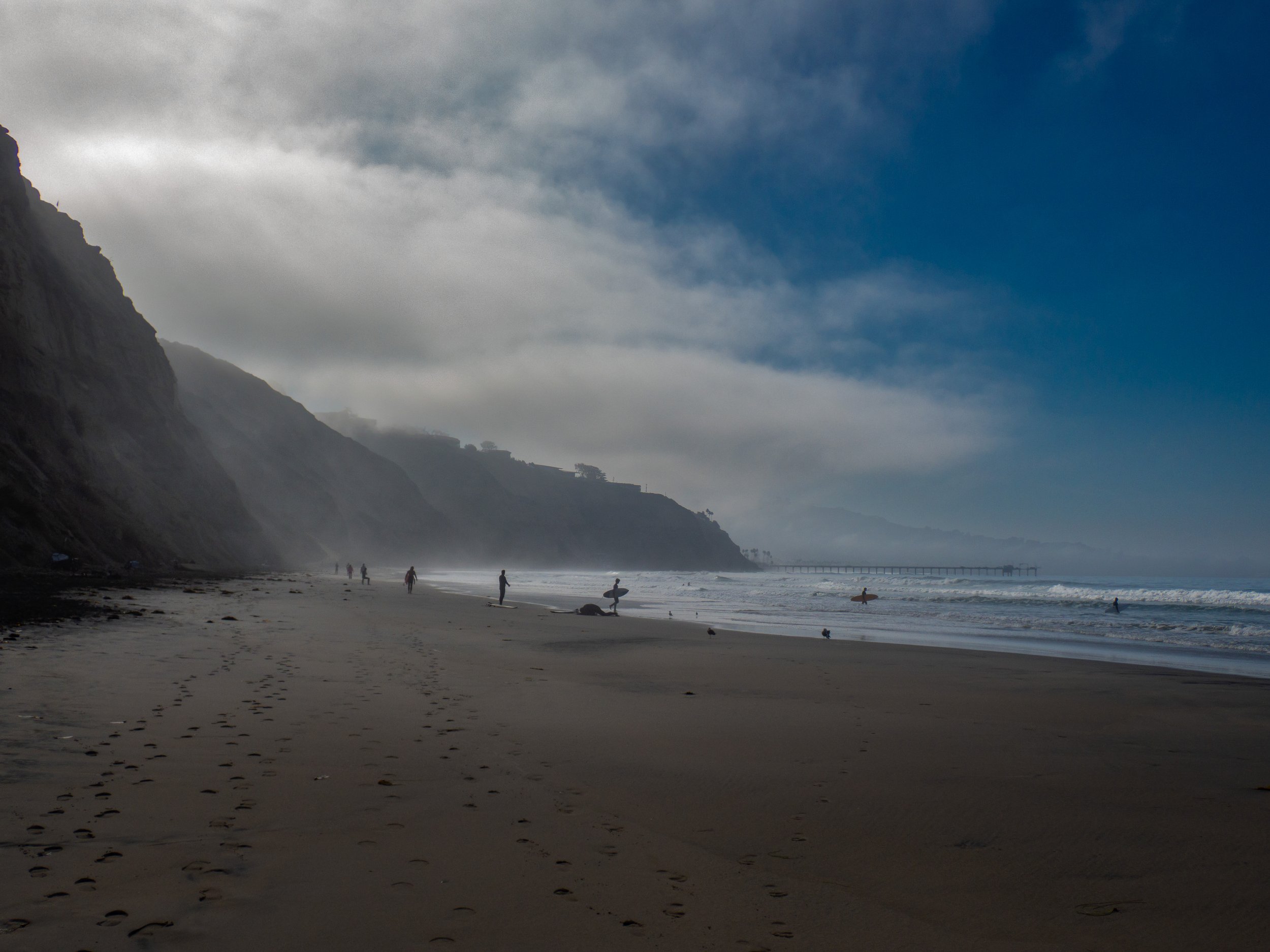 A beach with cliffs on the left side, a pier in the distance, and several people walking or surfing in the water under a partly cloudy sky.