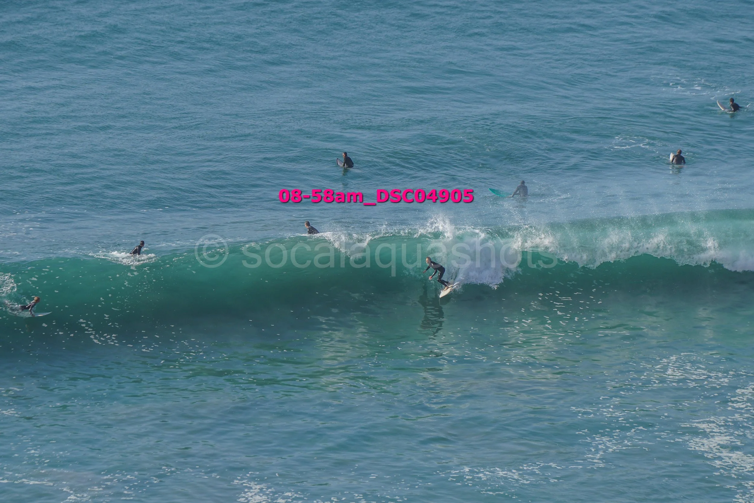 Surfers riding and waiting on ocean waves at the beach, some standing on their surfboards, others paddling in the water.