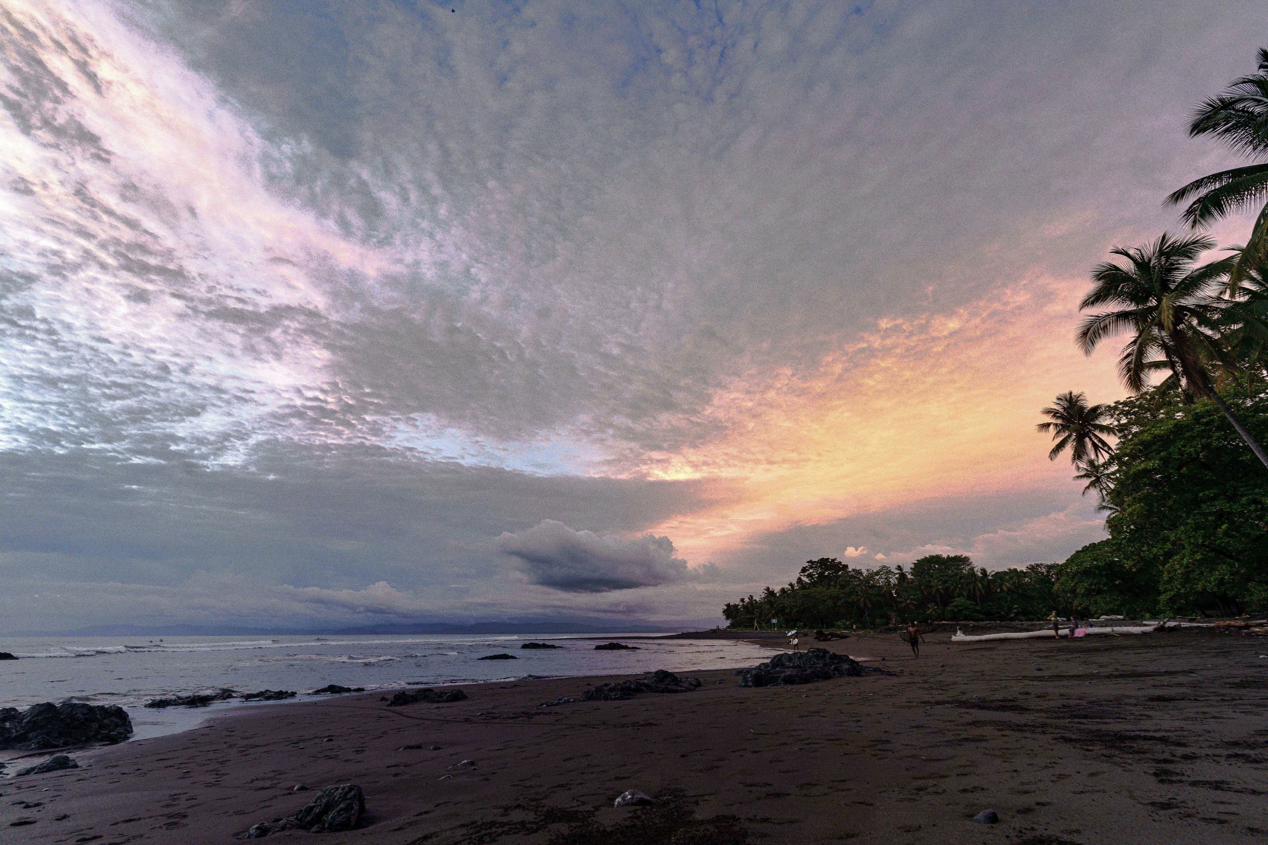 A beach scene at dusk with a colorful sunset sky, palm trees on the right, ocean waves, rocks on the shore, and a few people walking along the beach.