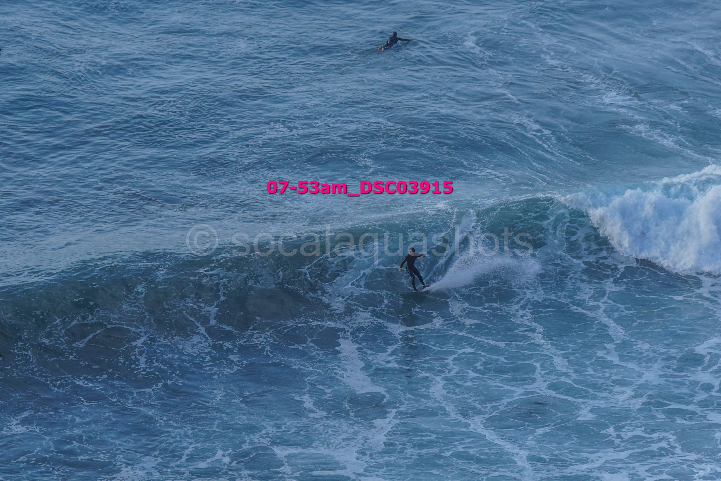 Surfer riding a wave in the ocean with another surfer visible farther away, water splashing around.