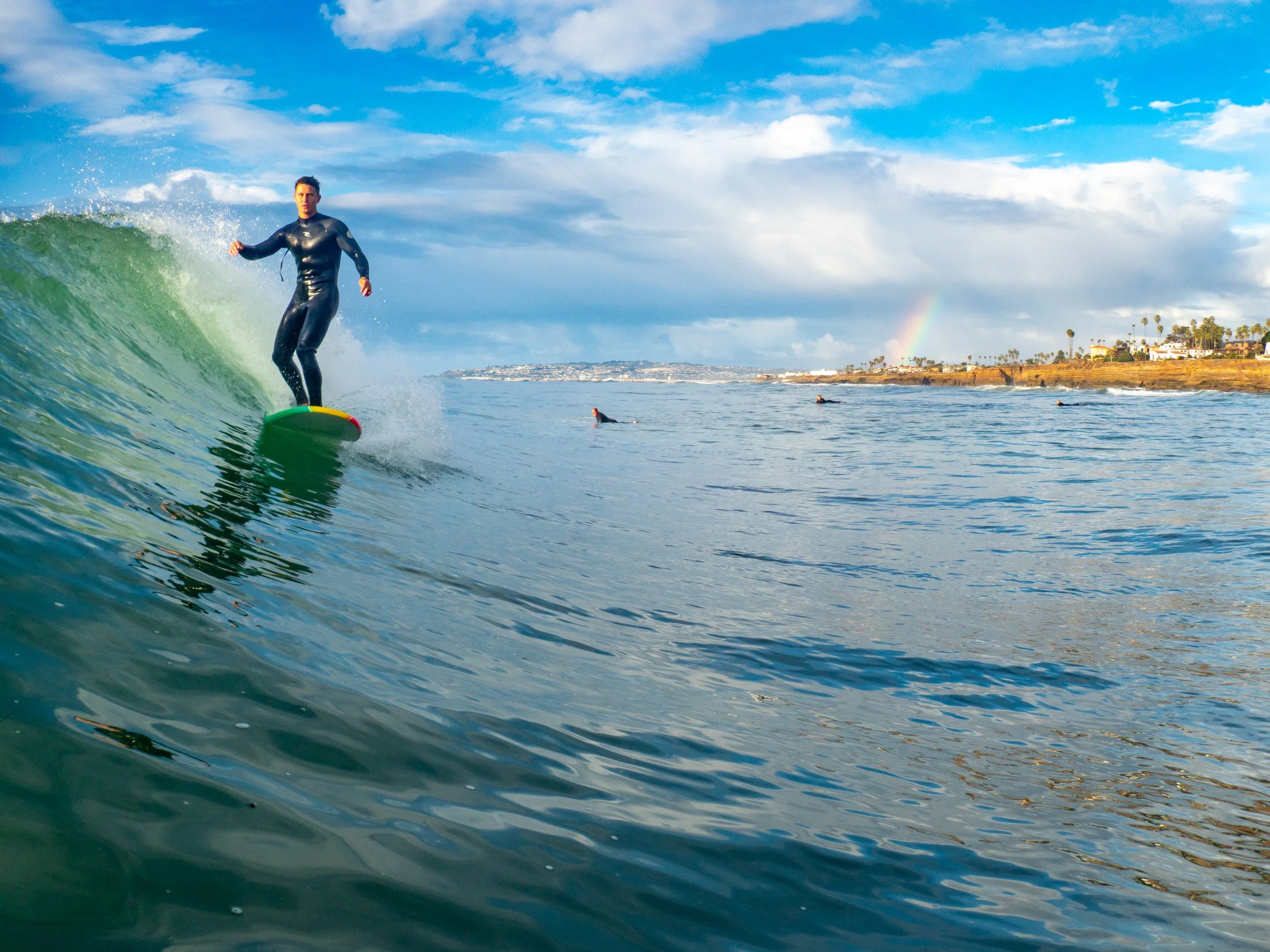 A person in a wetsuit surfing on a green surfboard in the ocean with a coastline, palm trees, a rainbow, and a partly cloudy sky in the background.
