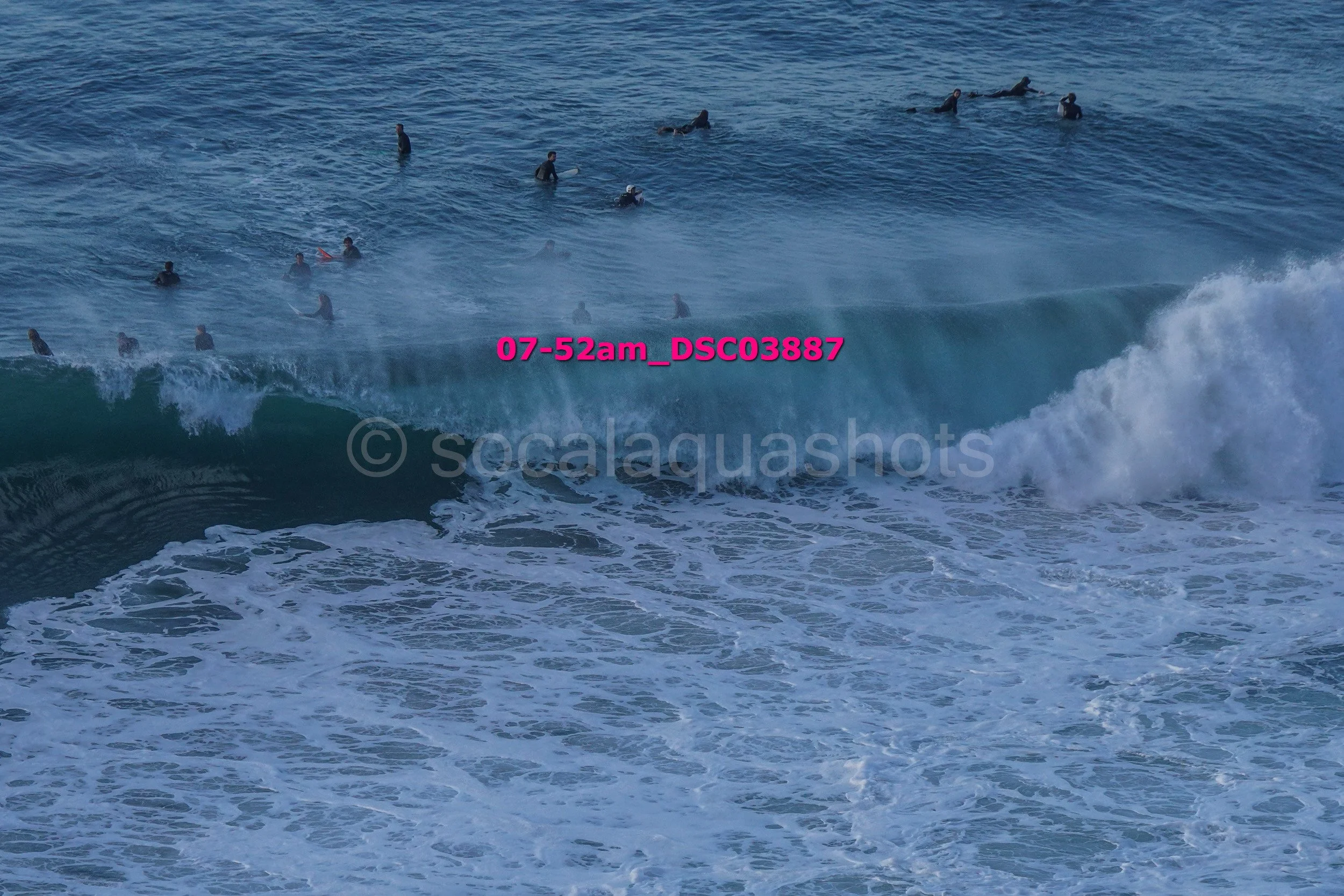  surfers in wetsuits in the ocean with waves crashing, some waiting and others riding the surf.