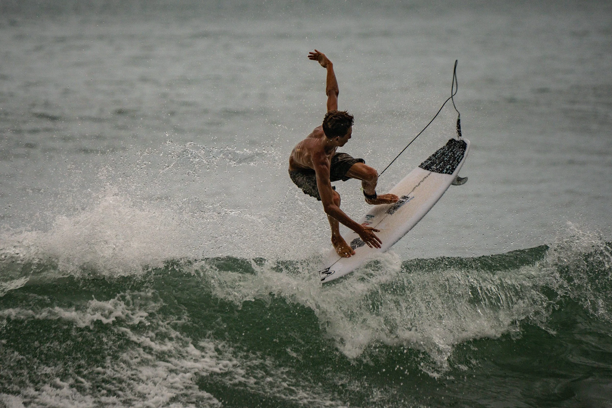 A person with short dark hair surfing on a white surfboard in the ocean, wearing dark board shorts, with an expression of focus as they balance on the wave.