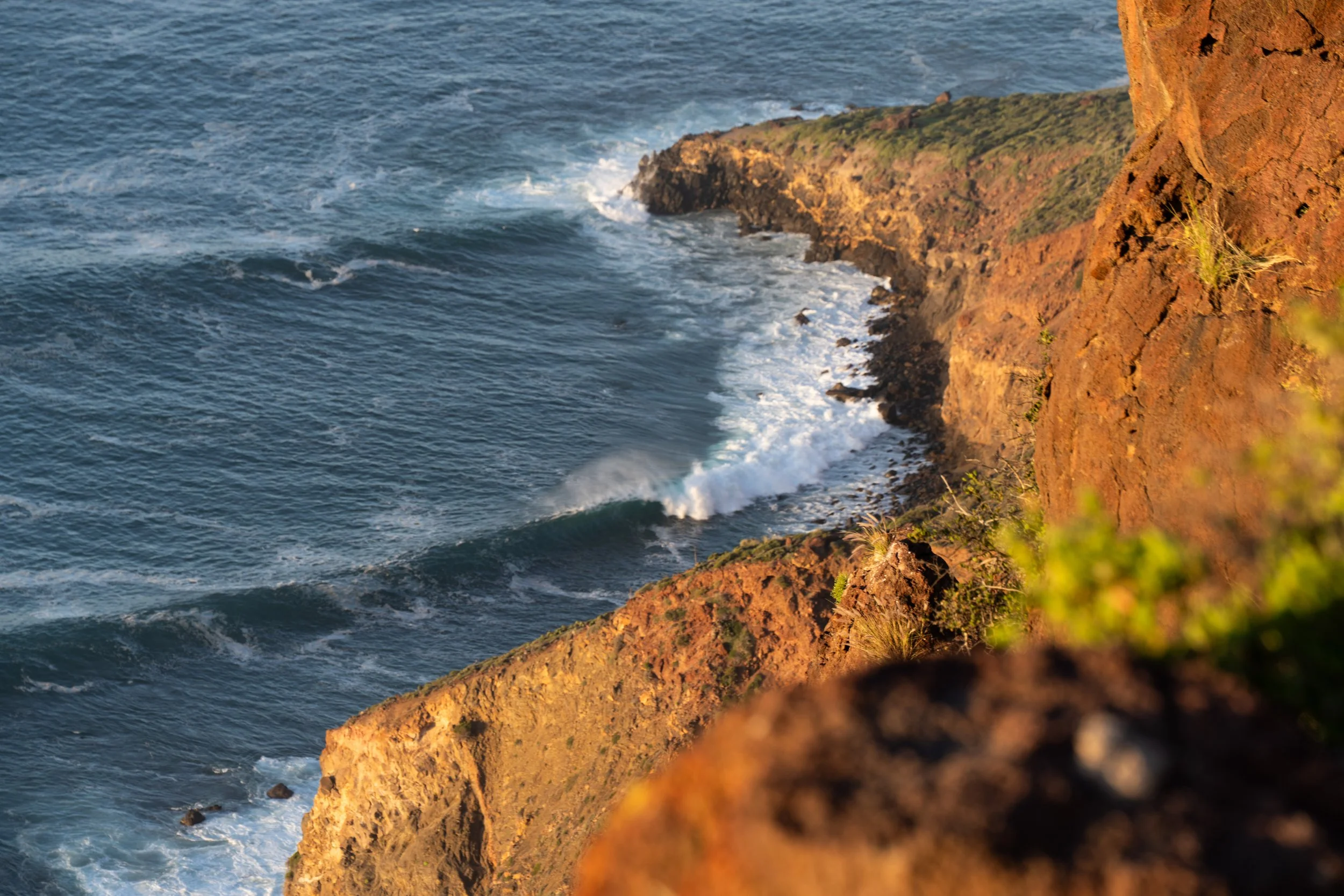 View of ocean waves crashing against rocky cliffs with sparse green vegetation at sunset.