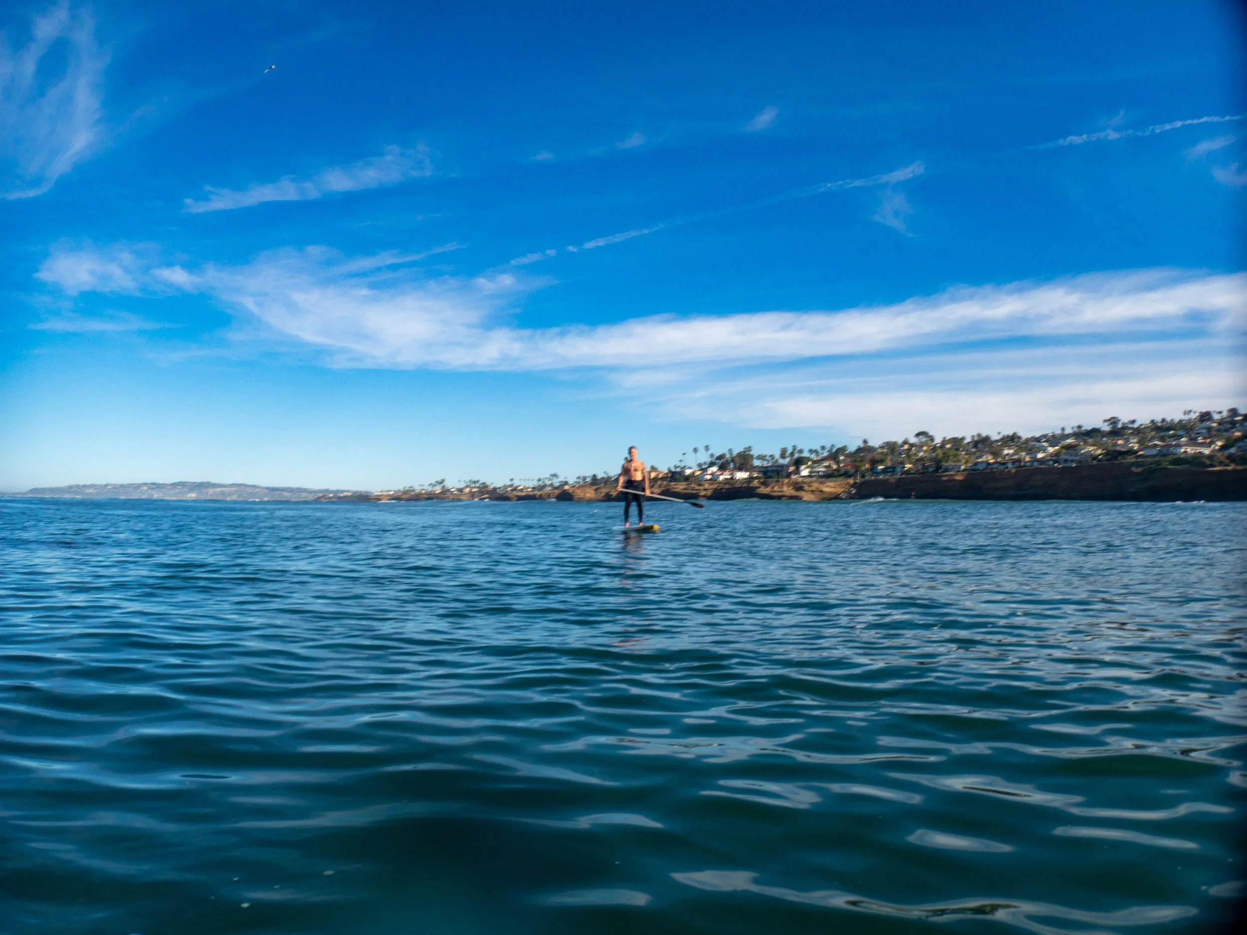 A person paddleboarding on calm ocean waters with a coastal town and clear blue sky in the background.