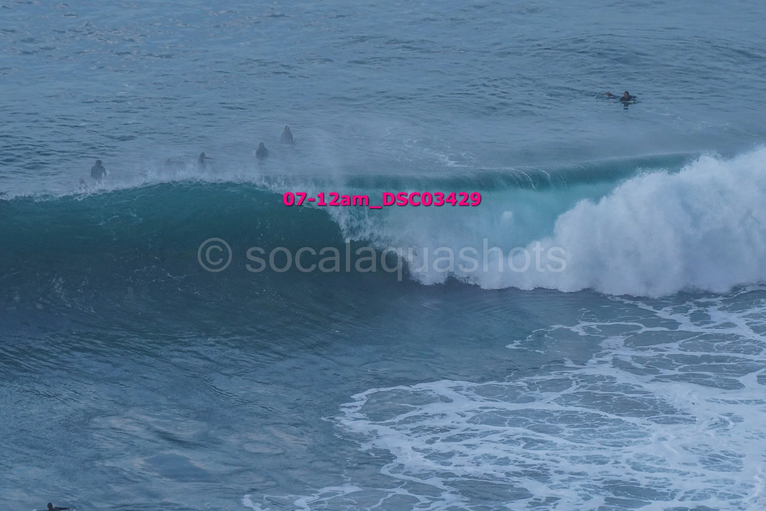 Surfers in the ocean riding a large wave, with some surfers visible in the water behind the wave.