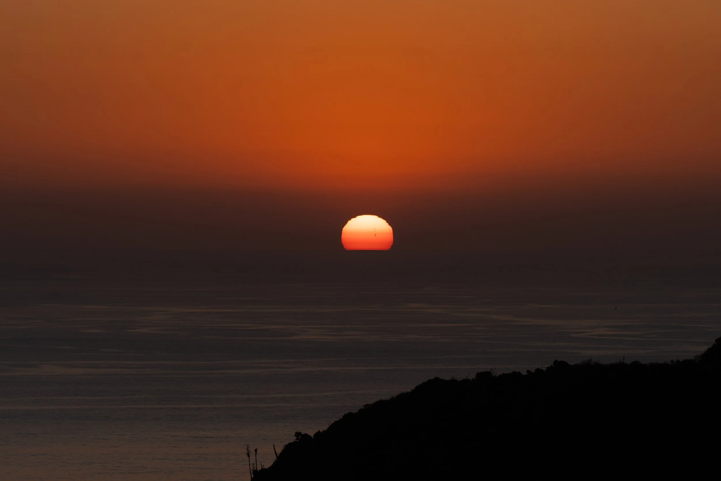 Sunset over the ocean with an orange and pink sky, a setting sun near the horizon, and a silhouette of a hill in the foreground.