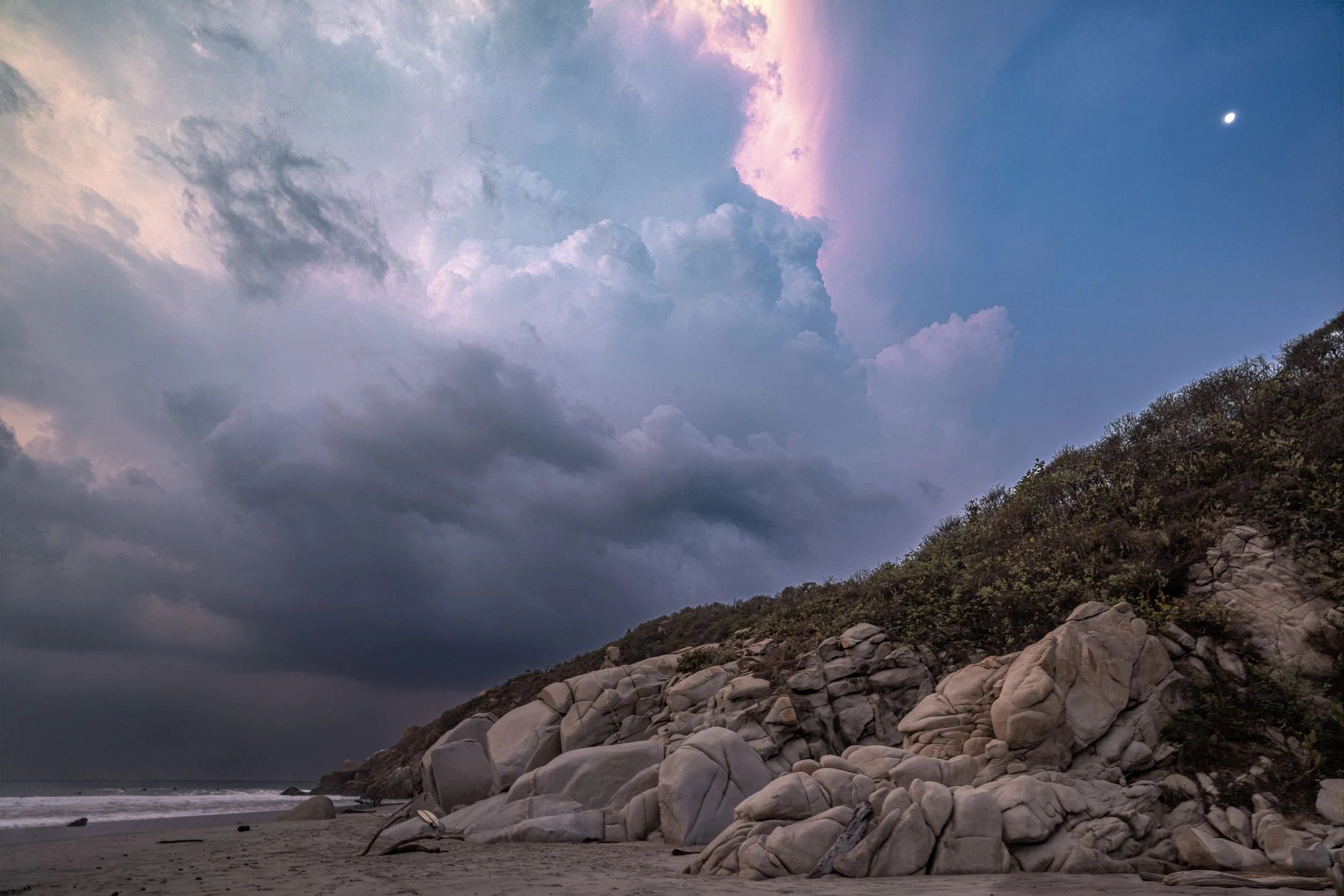 A landscape scene featuring a rocky shoreline with large stones and a hillside covered in shrubs. The sky is partly cloudy with dark clouds and some pink and purple hues, indicating sunset or sunrise, and a bright moon is visible in the blue sky.