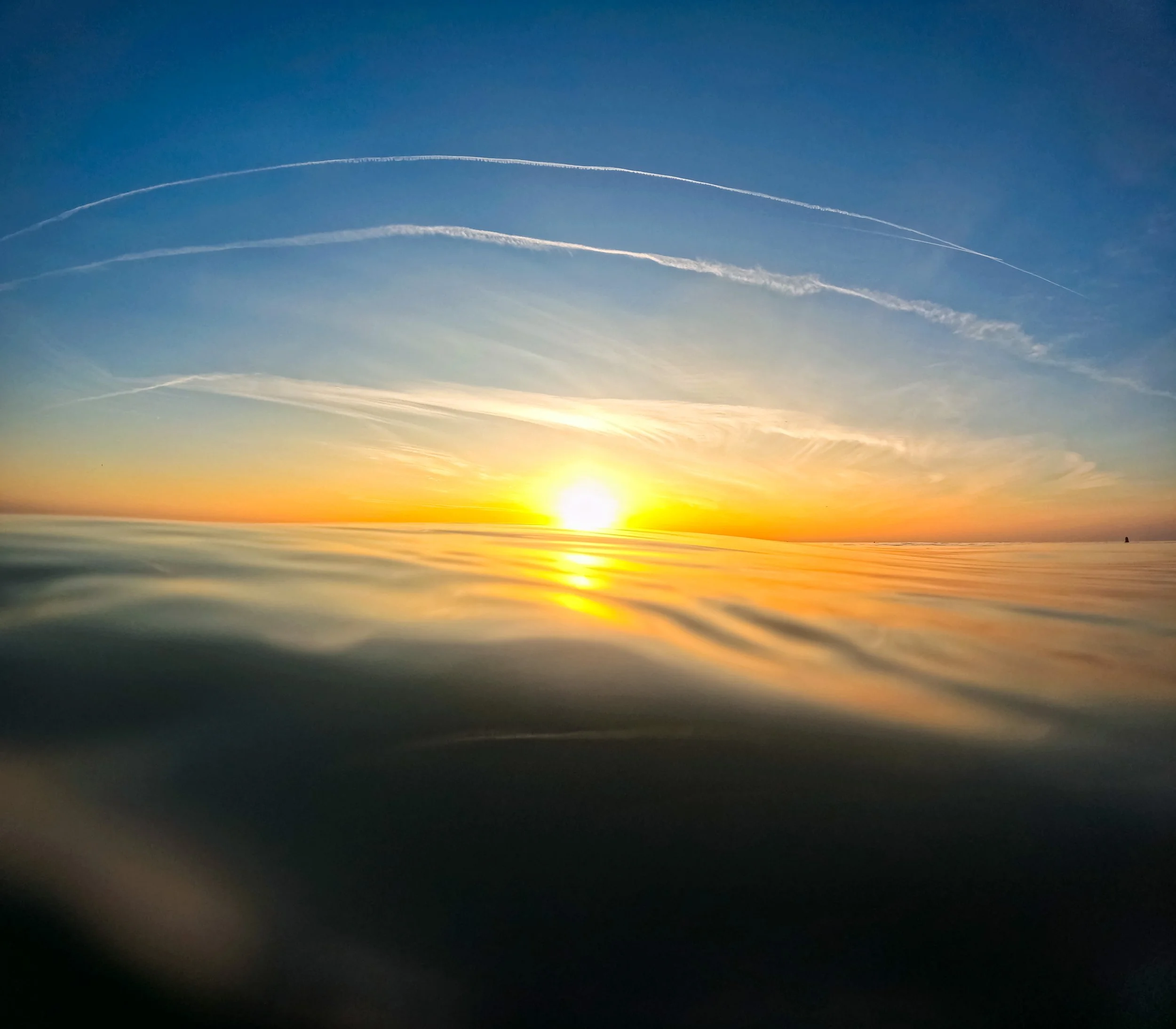 Sunset over the ocean with the sun low on the horizon, the sky is blue with streaks of white clouds, and the water reflects the warm colors of the sunset.