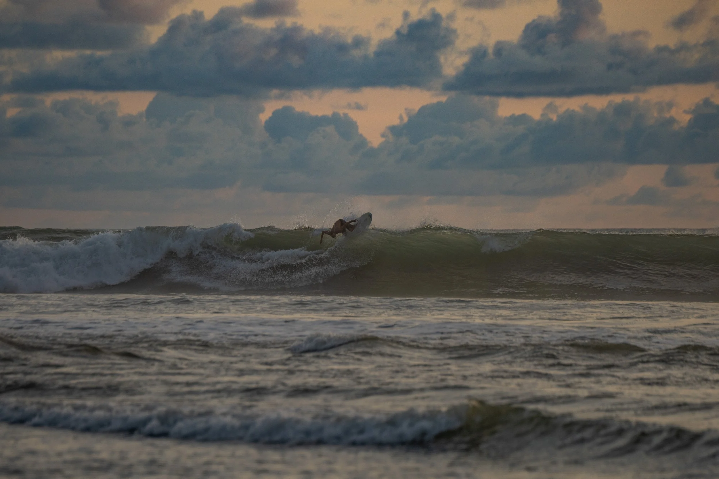 A surfer riding a wave in the ocean during sunset or sunrise under a cloudy sky.