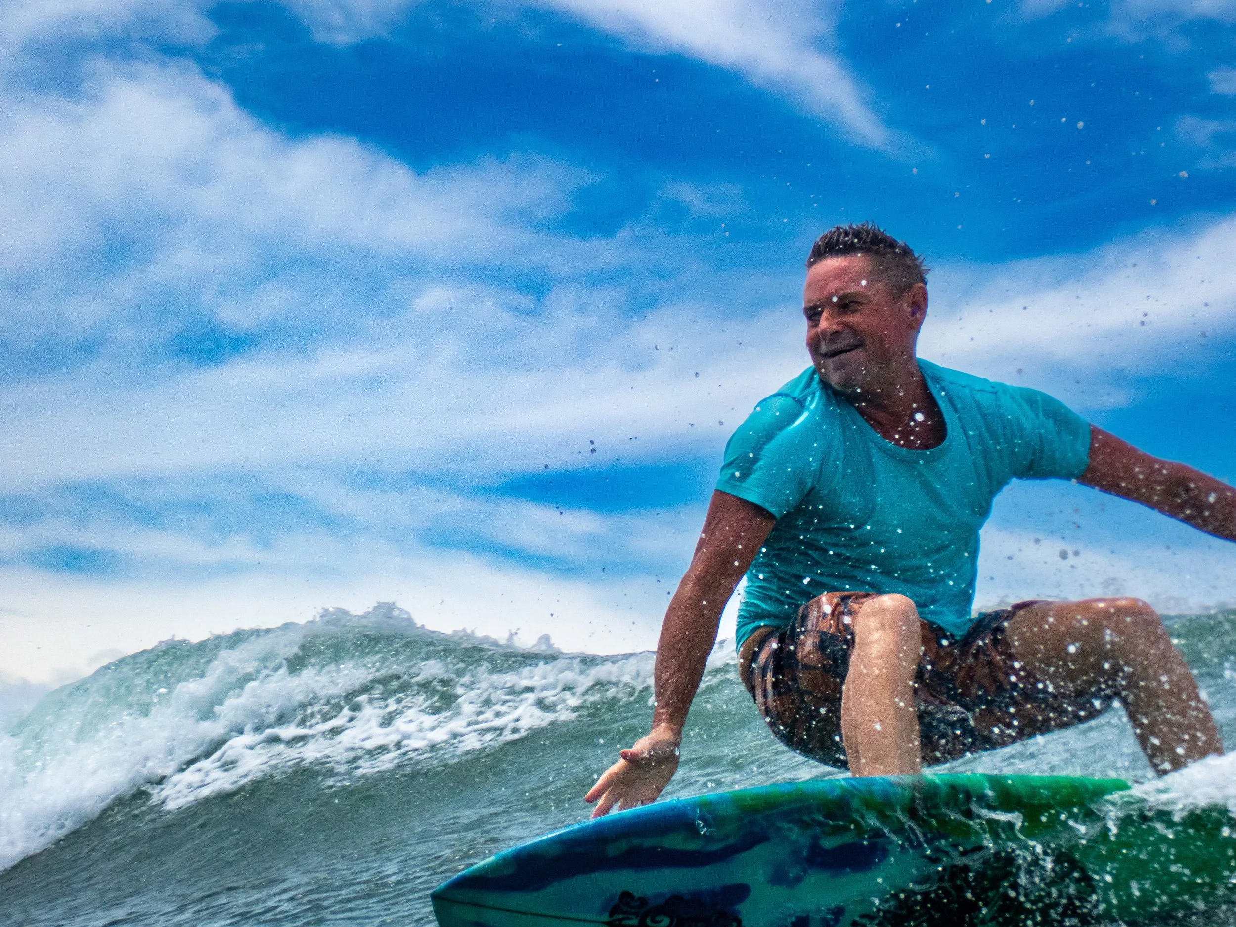 A man surfing in the ocean with a blue sky in the background, wearing a blue shirt and dark board shorts, riding a small wave.