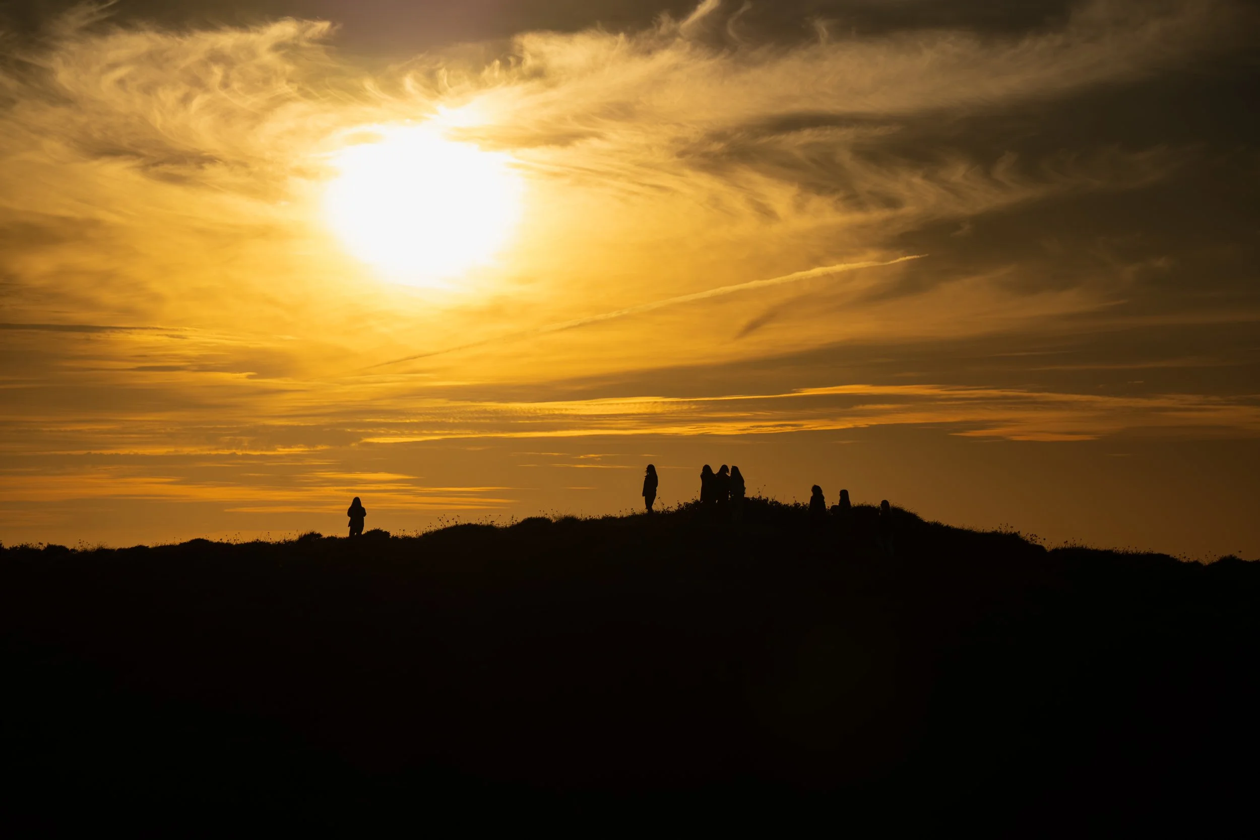 Silhouette of people on a hilltop during sunset with a golden sky and wispy clouds.