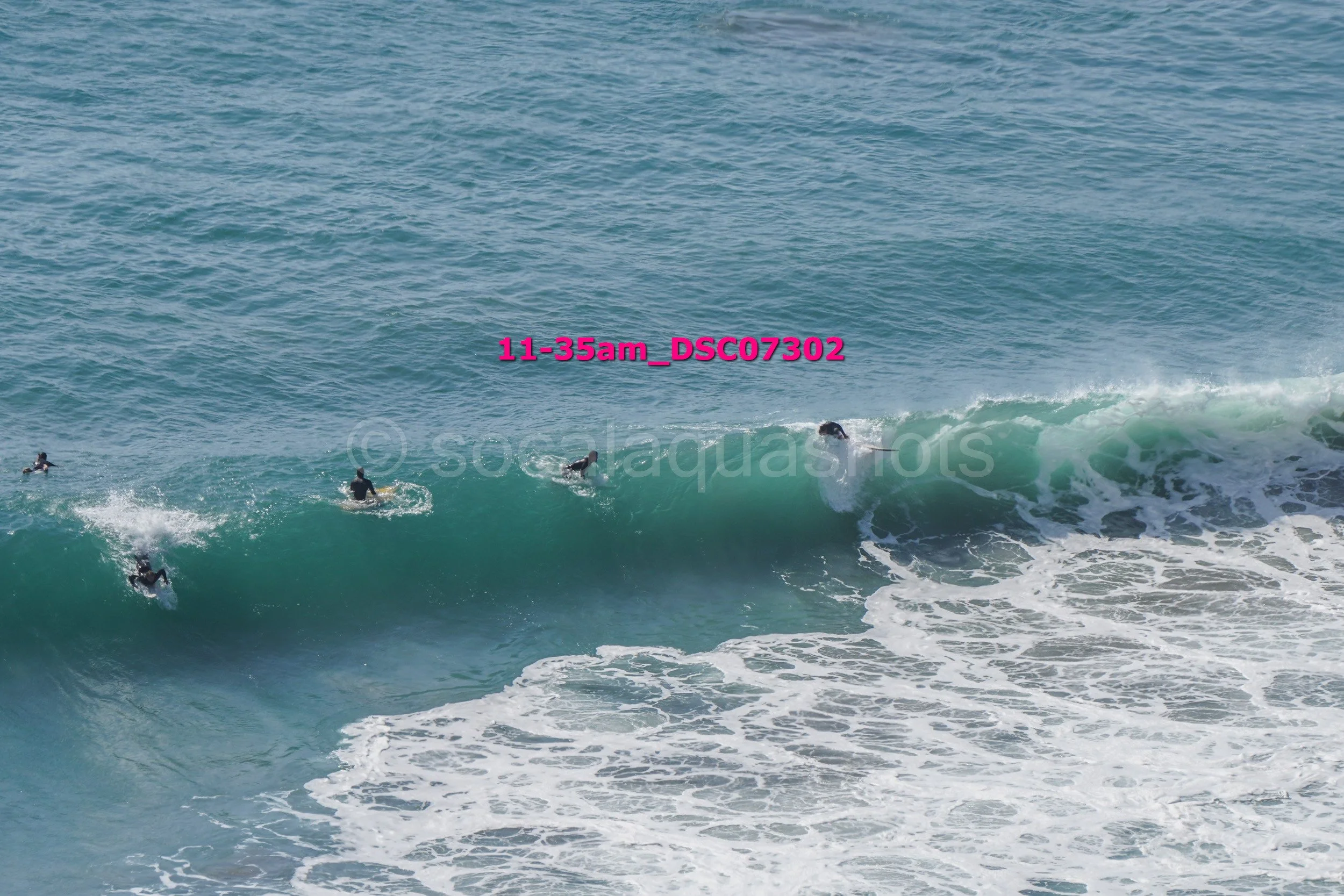 Four surfers in wetsuits riding a wave in the ocean, with one wave curling over and white foam at the bottom.