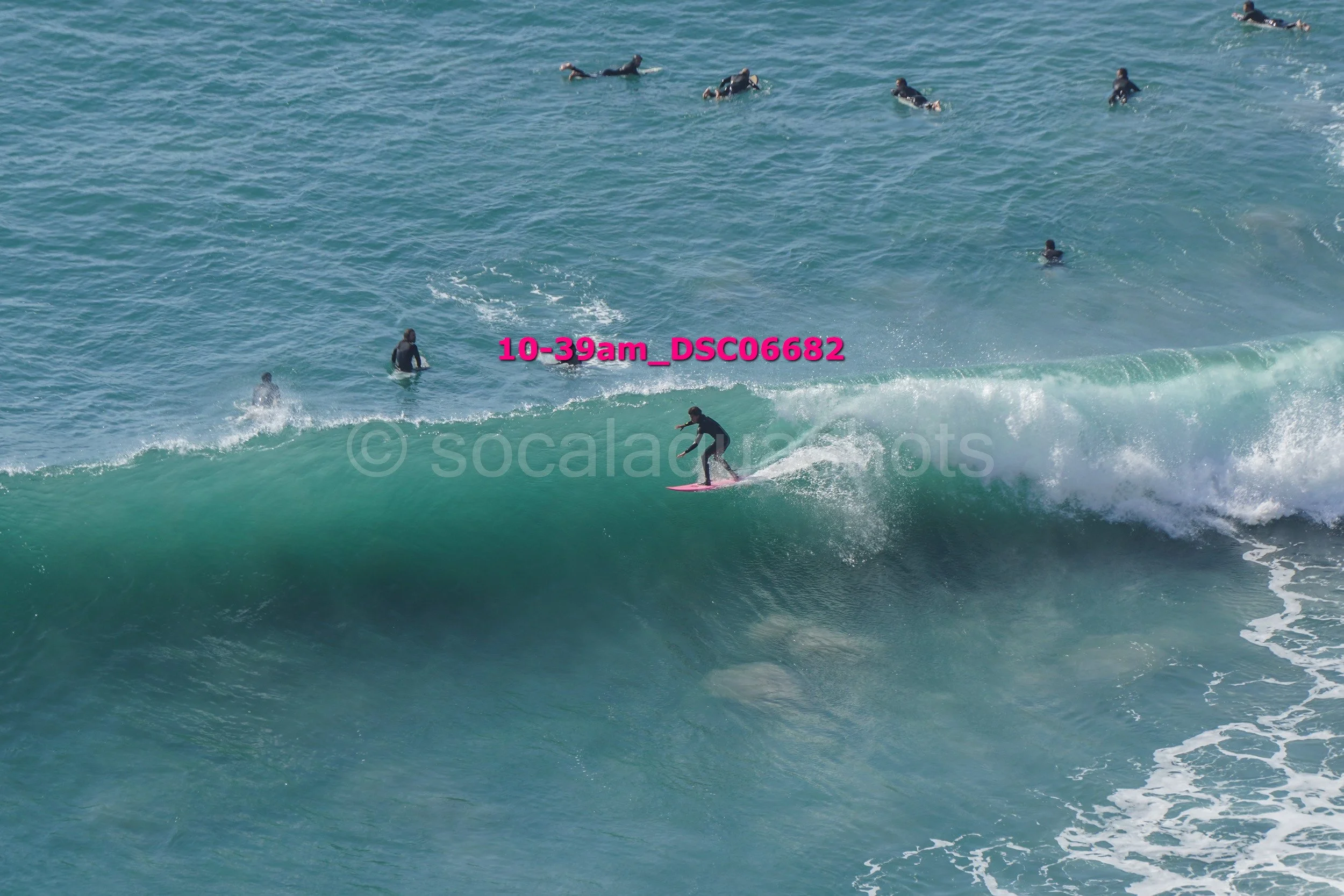 A person riding a pink surfboard on a large wave with several other people in the background in the water.