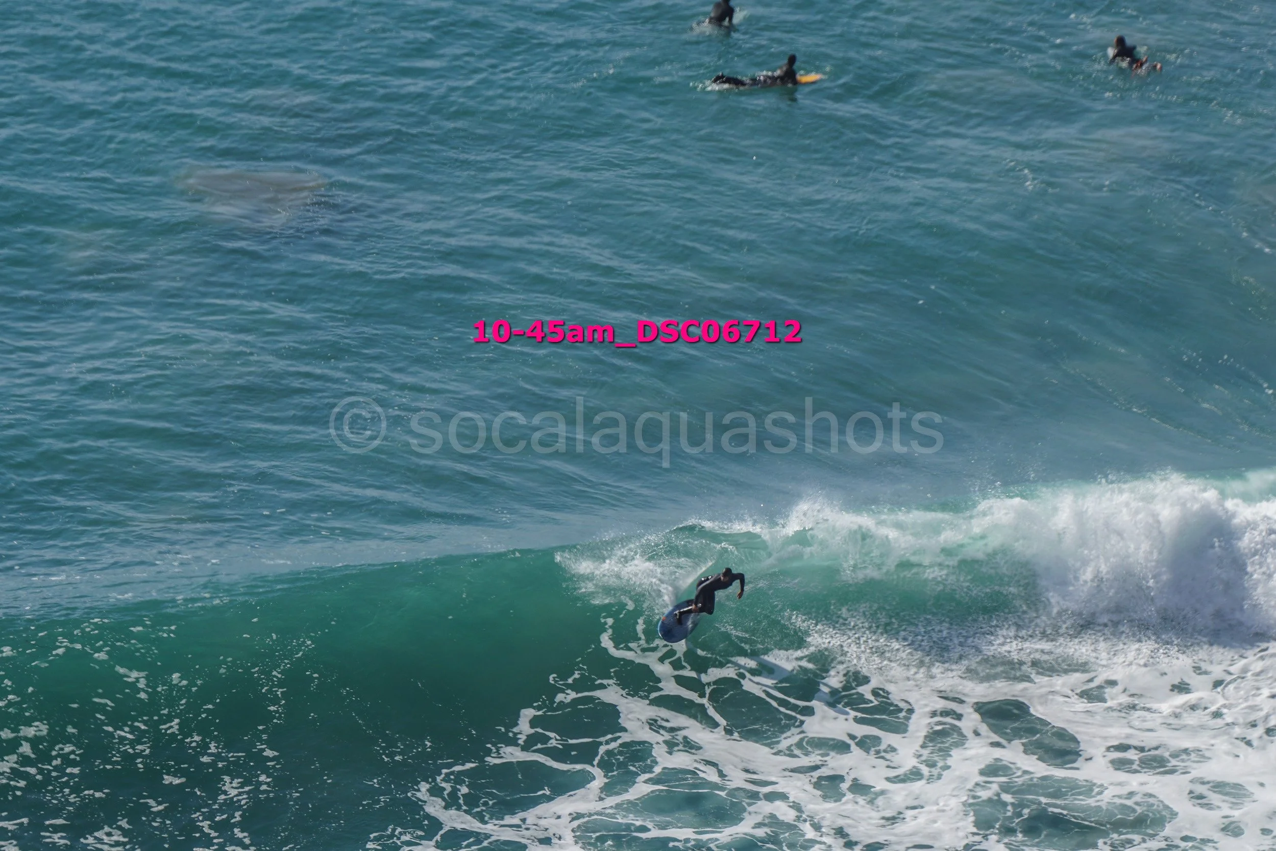 A surfer in a wetsuit riding a wave near the shoreline with several other surfers visible in the water in the background.