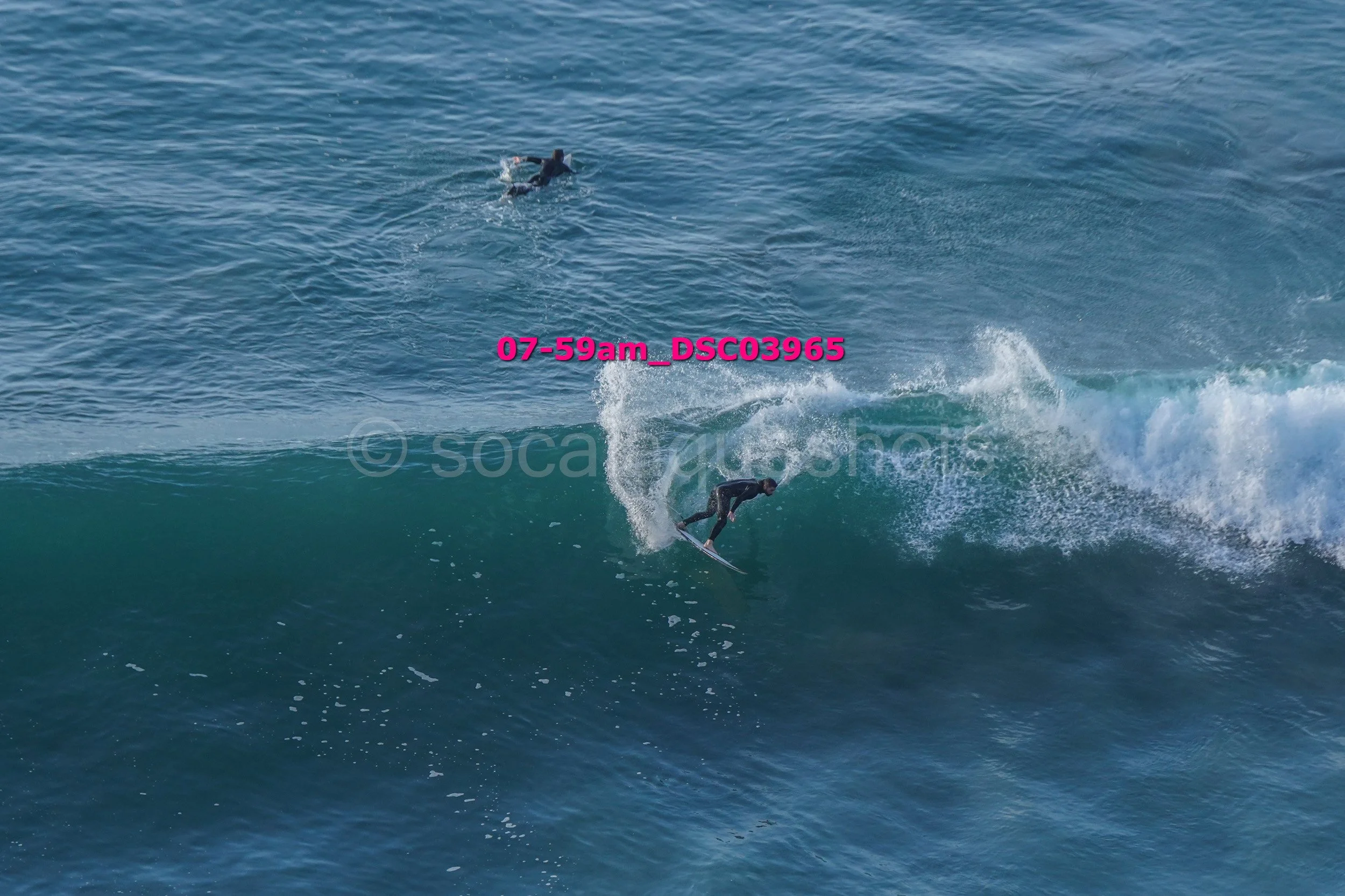 A person riding a wave on a surfboard in the ocean with another surfer visible in the background and pink text overlay.