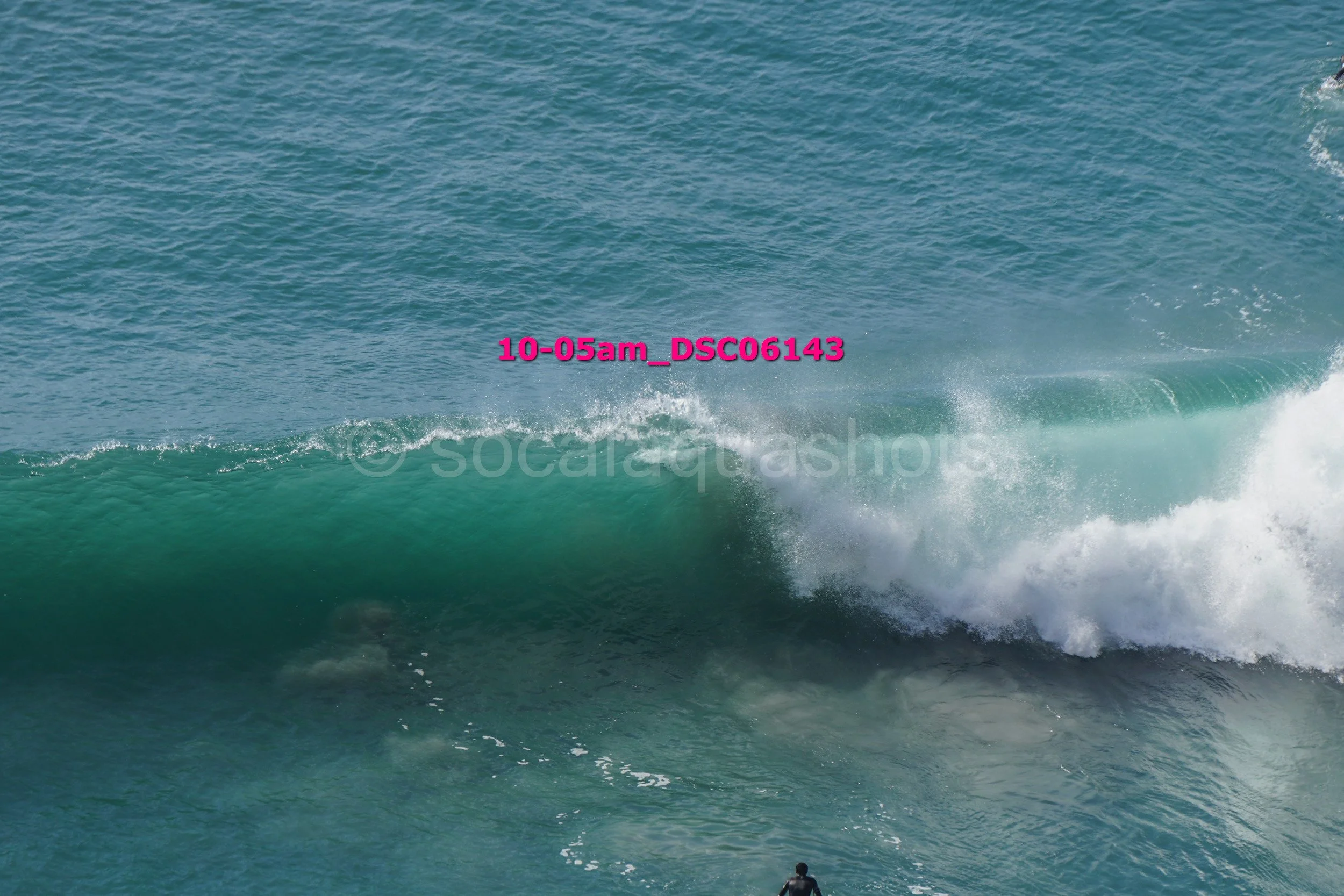 Large ocean wave with white foam, viewed from below. A person in a wetsuit stands in the water at the bottom of the wave.