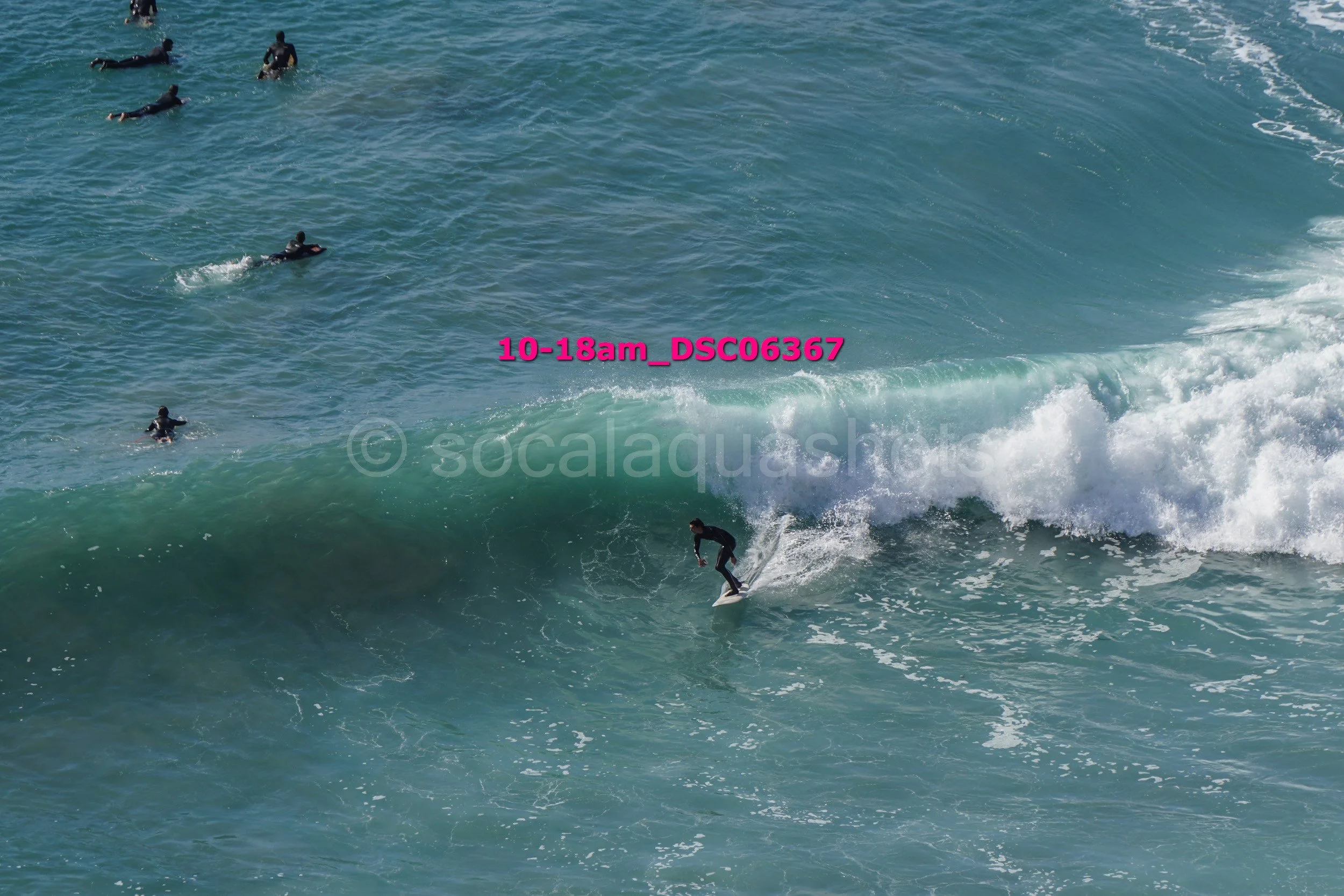Surfer riding a wave in the ocean while several other surfers float on their boards nearby.
