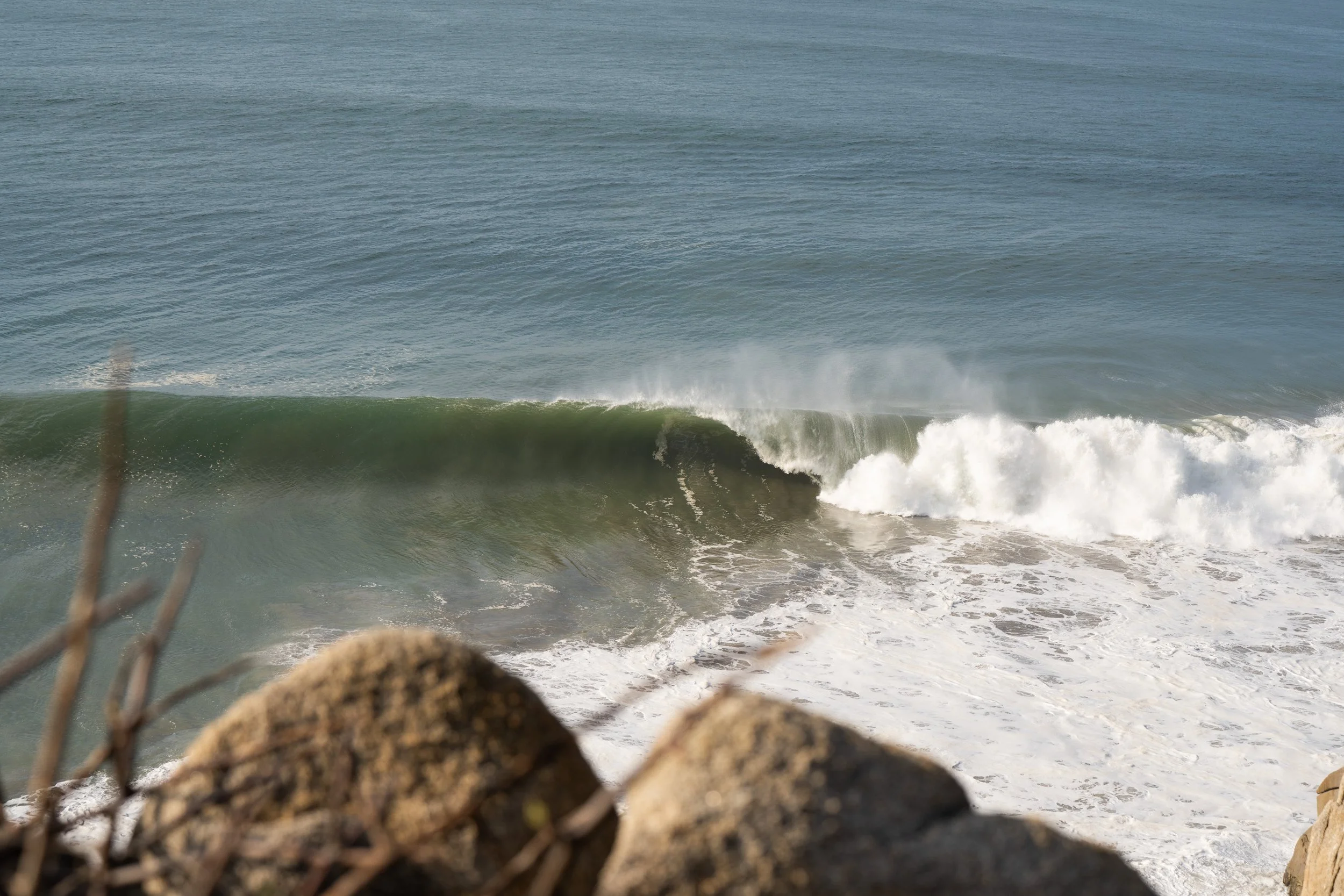 Ocean waves crashing against rocks along the shoreline, viewed through a chain-link fence with some blurred rocks in the foreground.