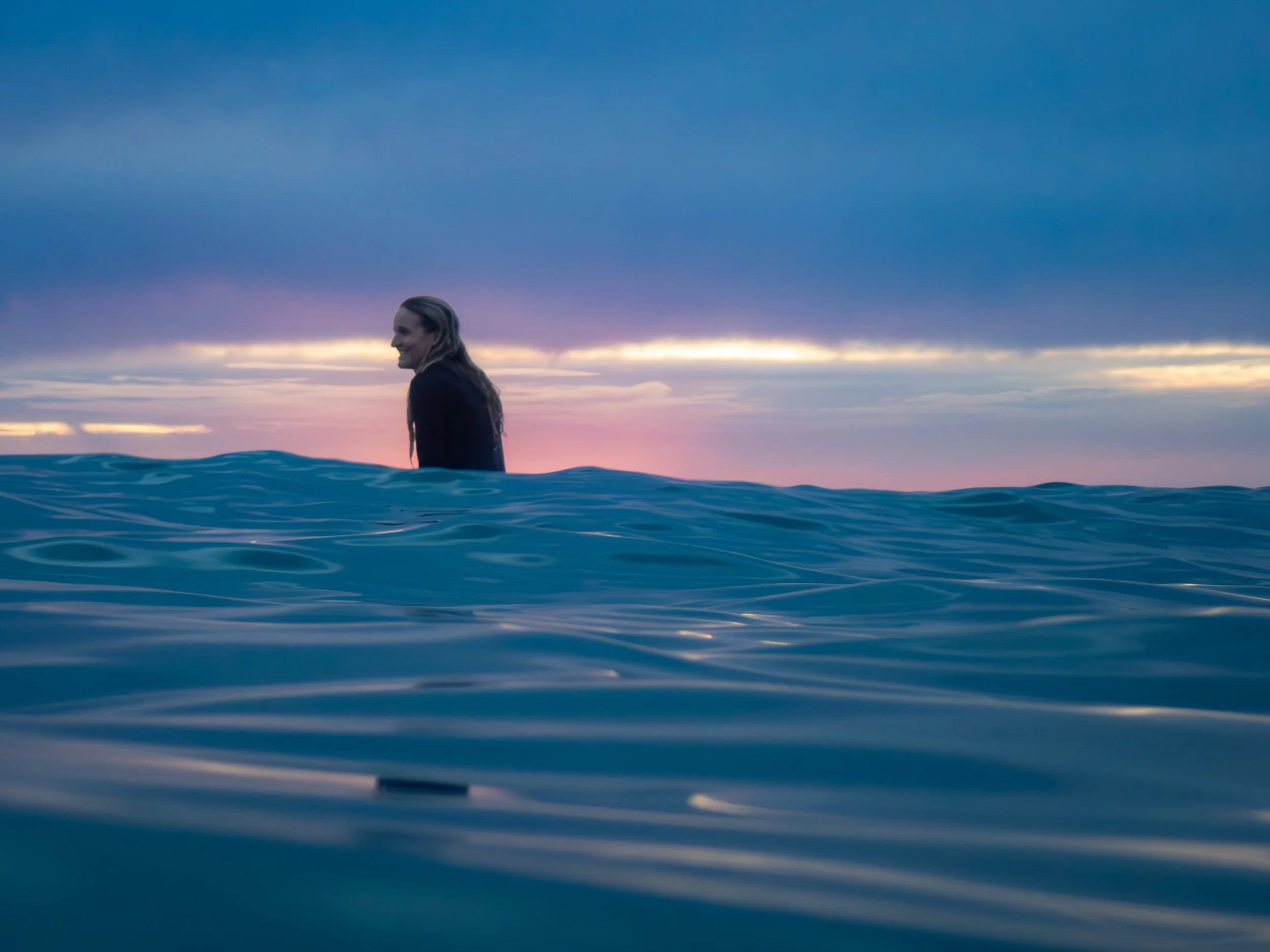A person in a wetsuit smiling in the ocean at sunset with a colorful sky and clouds.
