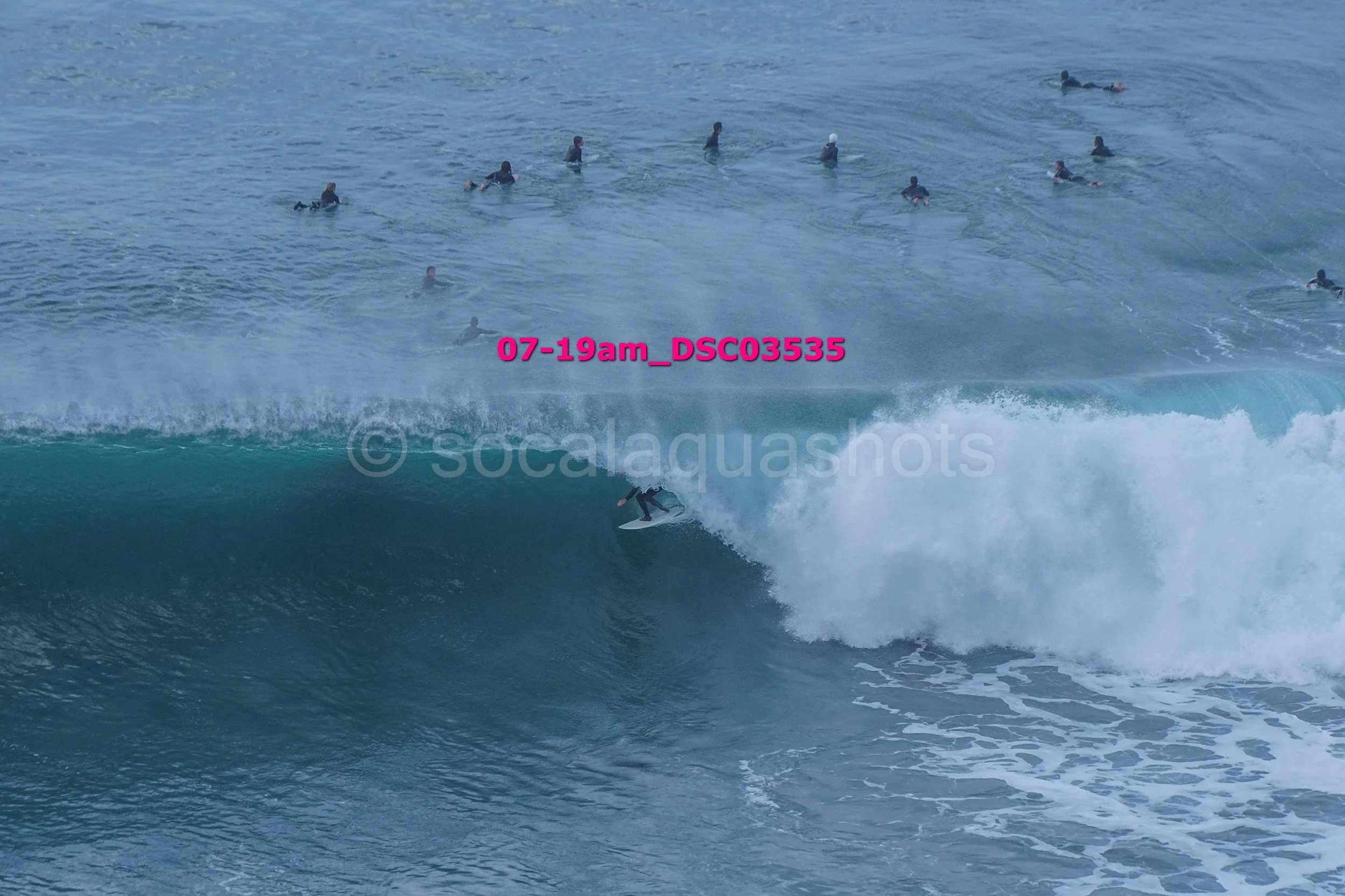 A person surfing on a wave with a large group of swimmers in the water nearby.