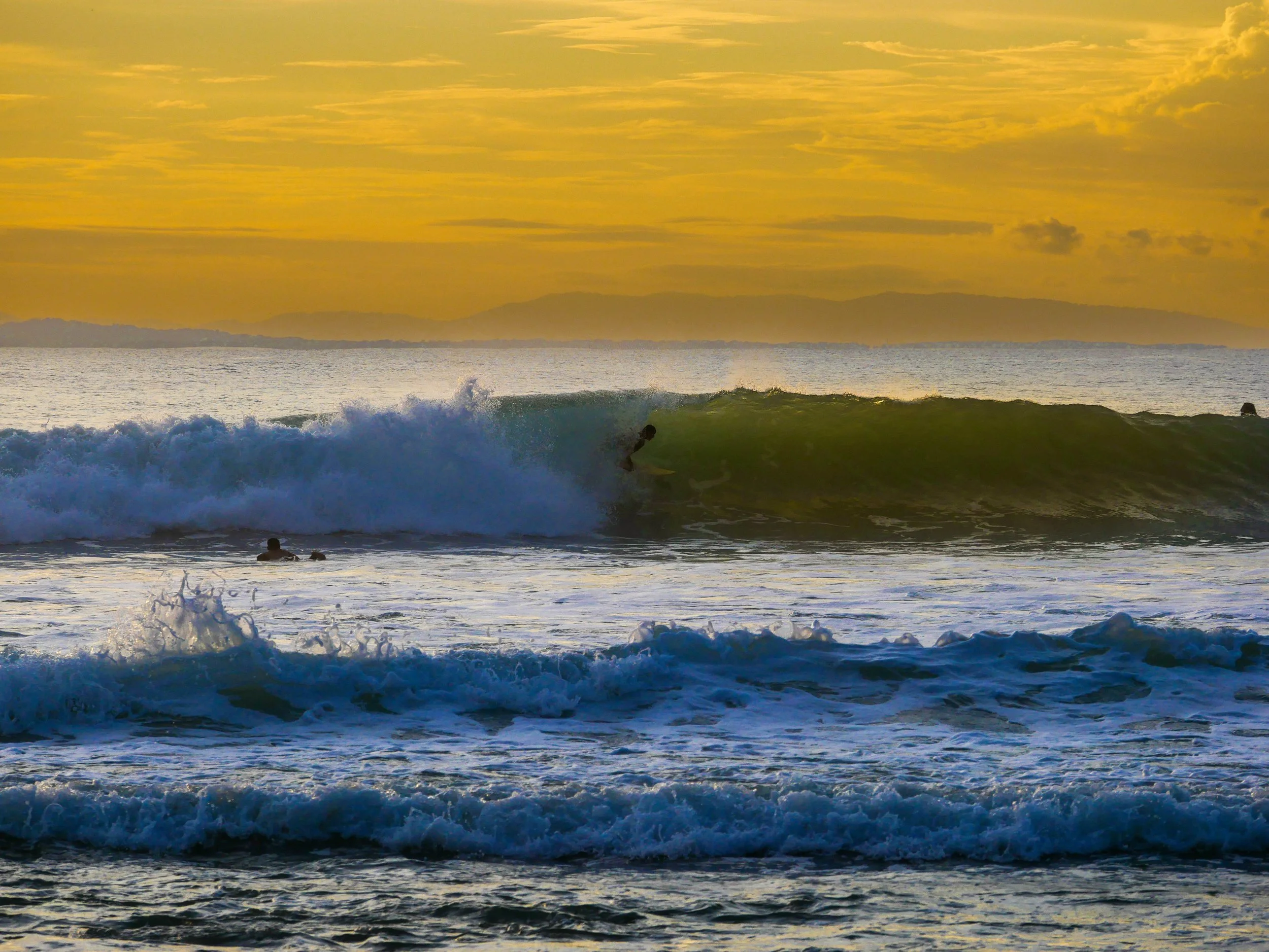 Surfer riding a wave at sunset with orange sky and distant hills.