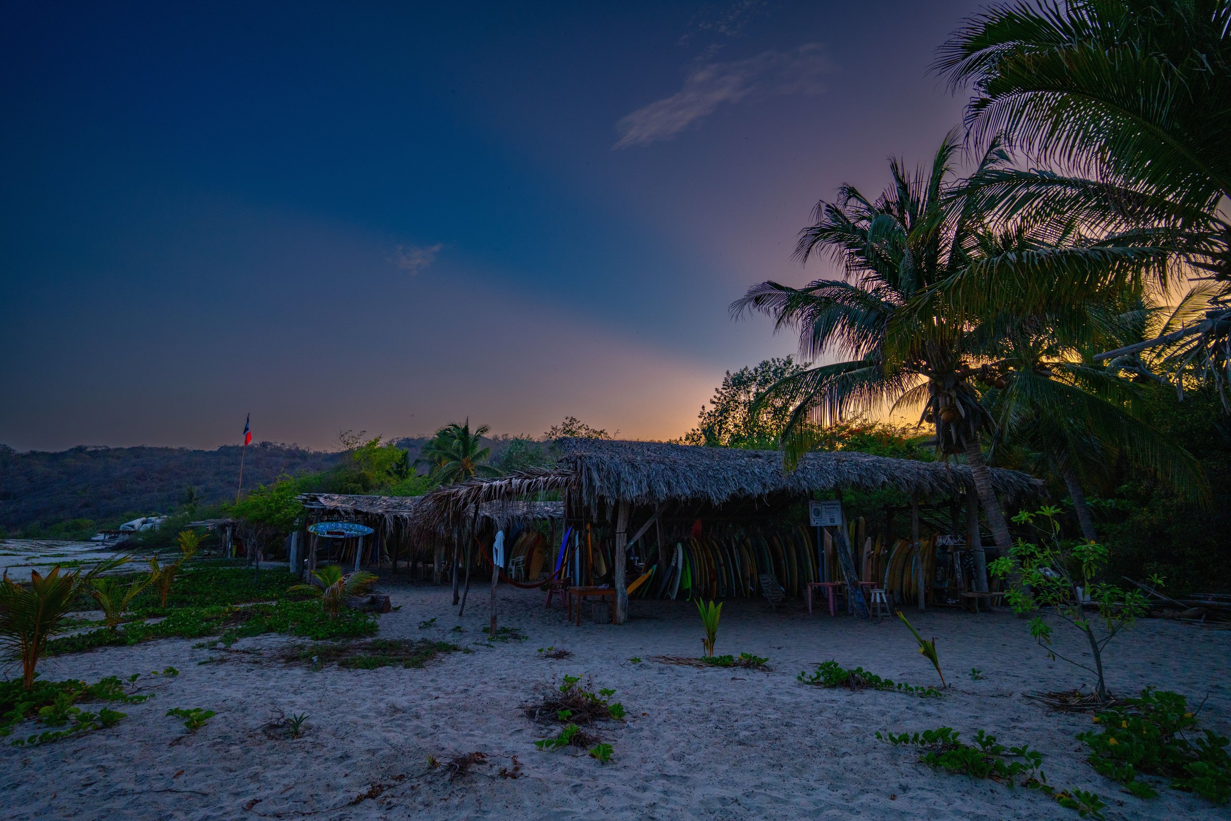 A sandy beach at sunset with palm trees and huts with thatched roofs. A flag is visible on the horizon, and surfboards are lined up under the huts.