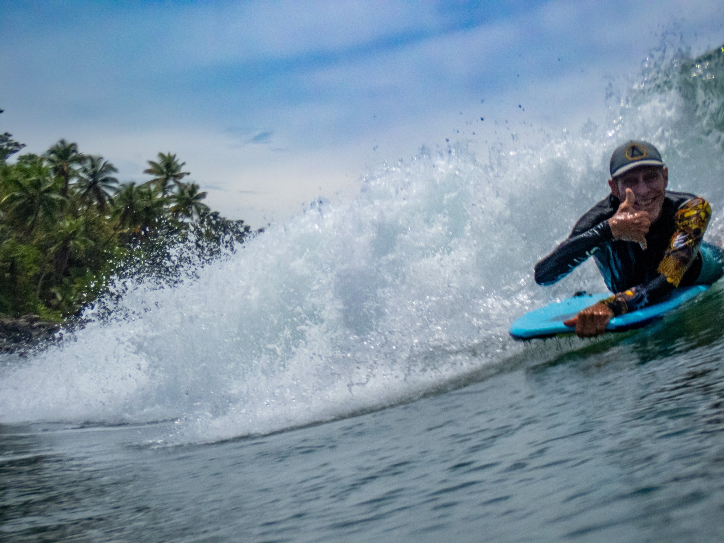 Person smiling and giving a thumbs-up while surfing on a blue surfboard in the ocean.