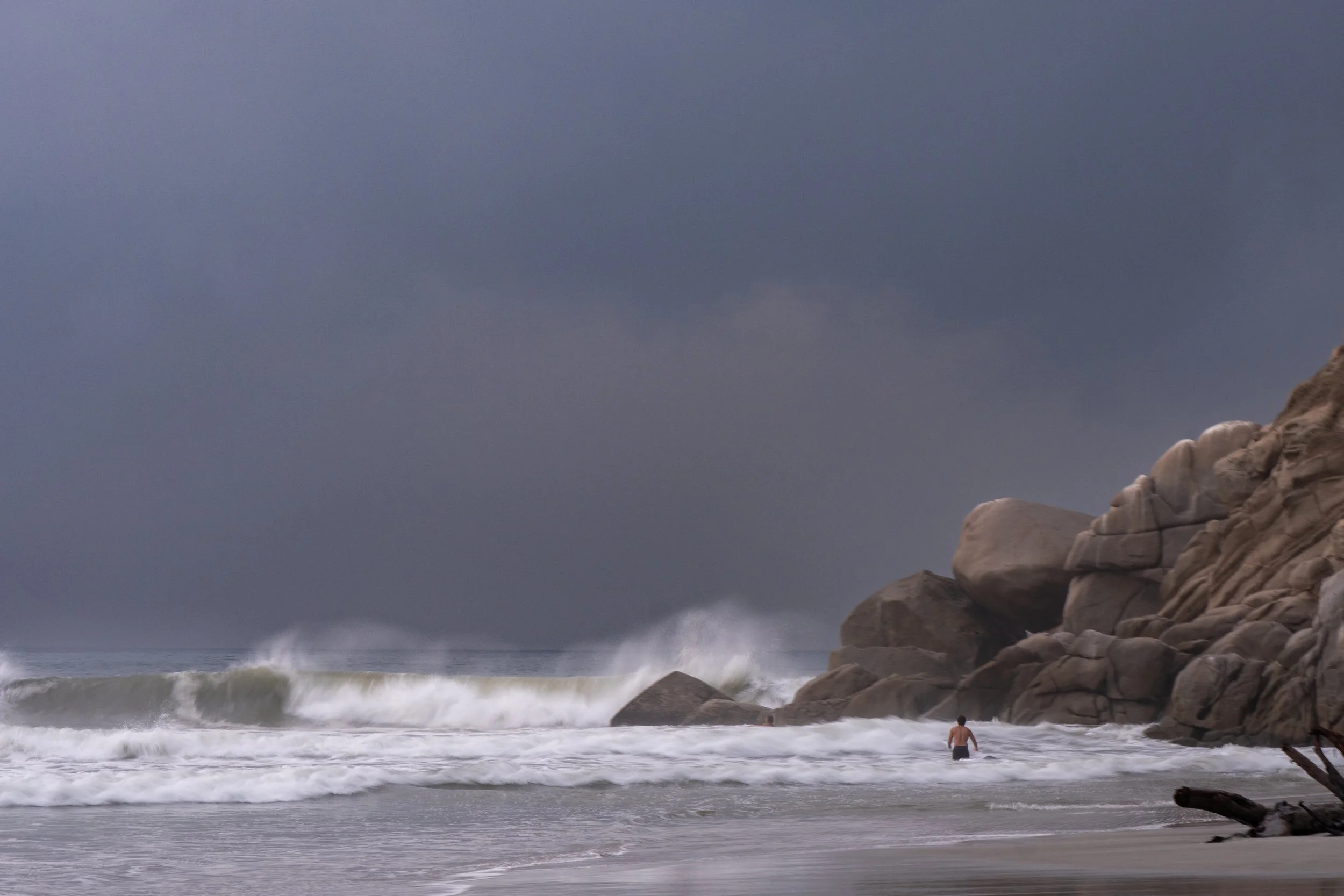 A person wading in the ocean near rocks on a cloudy day with large waves crashing against the rocks.
