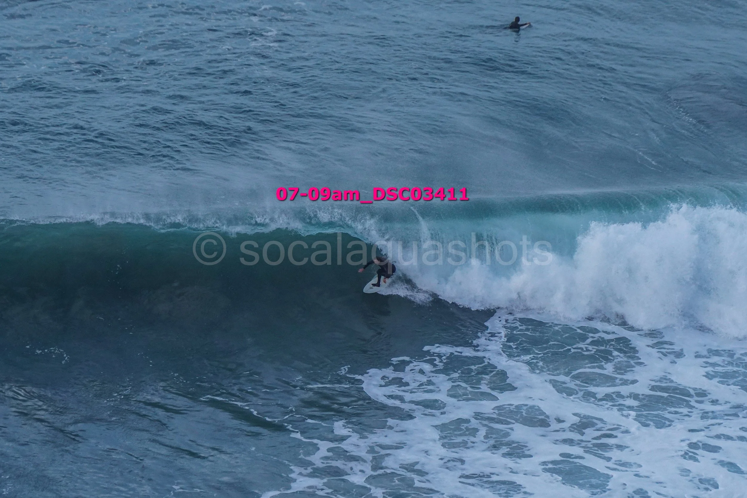 A surfer riding a large wave in the ocean, with another person on a surfboard visible in the background.